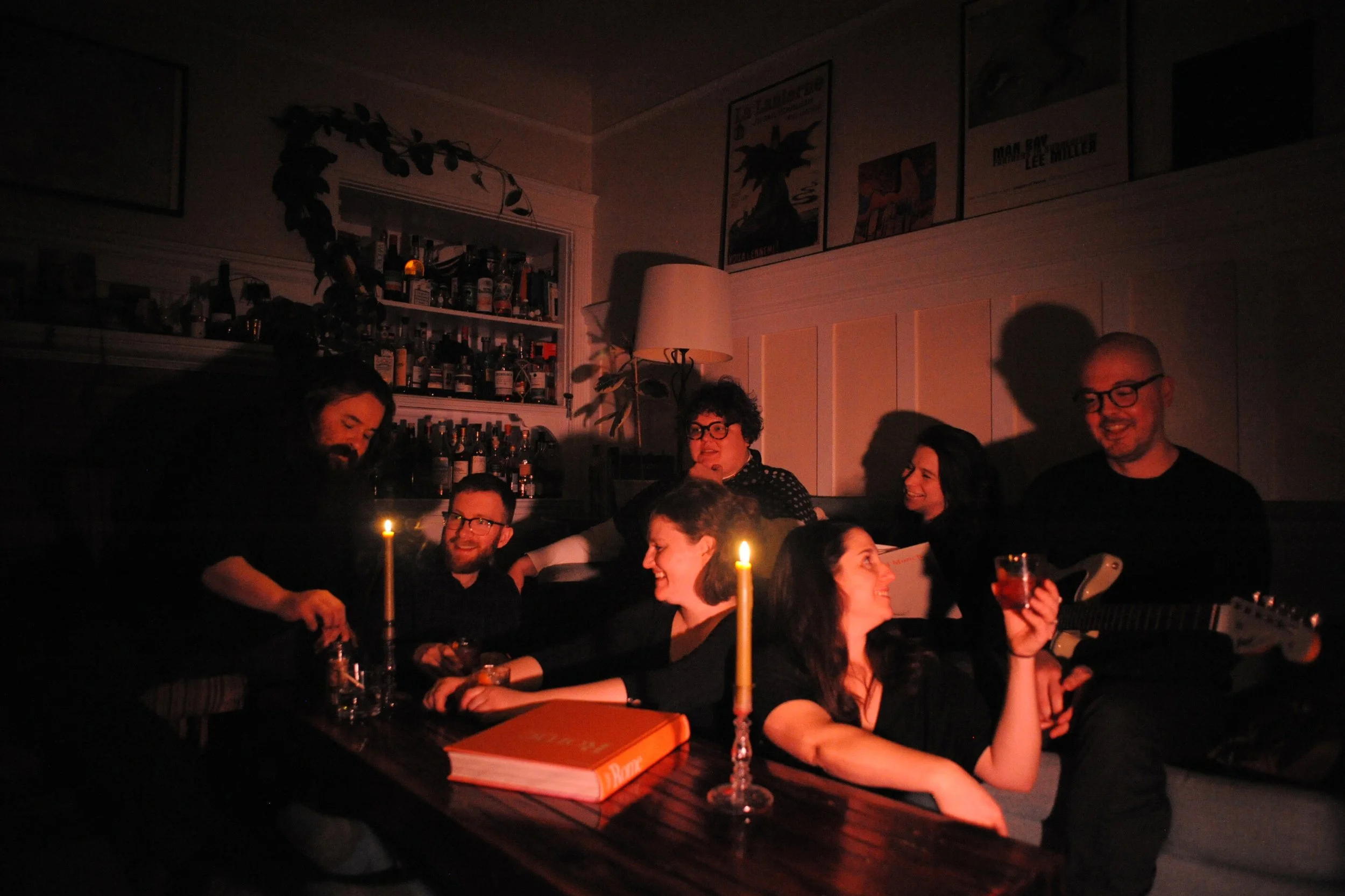 Group of people gathering in a dimly lit room, smiling and engaging with each other, with candles on the table and a bar in the background.