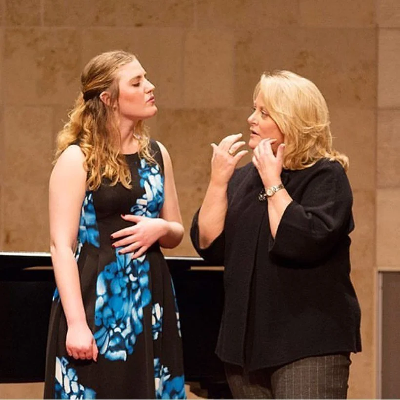 Two women engaged in a serious conversation in a music venue, with a grand piano in the background.