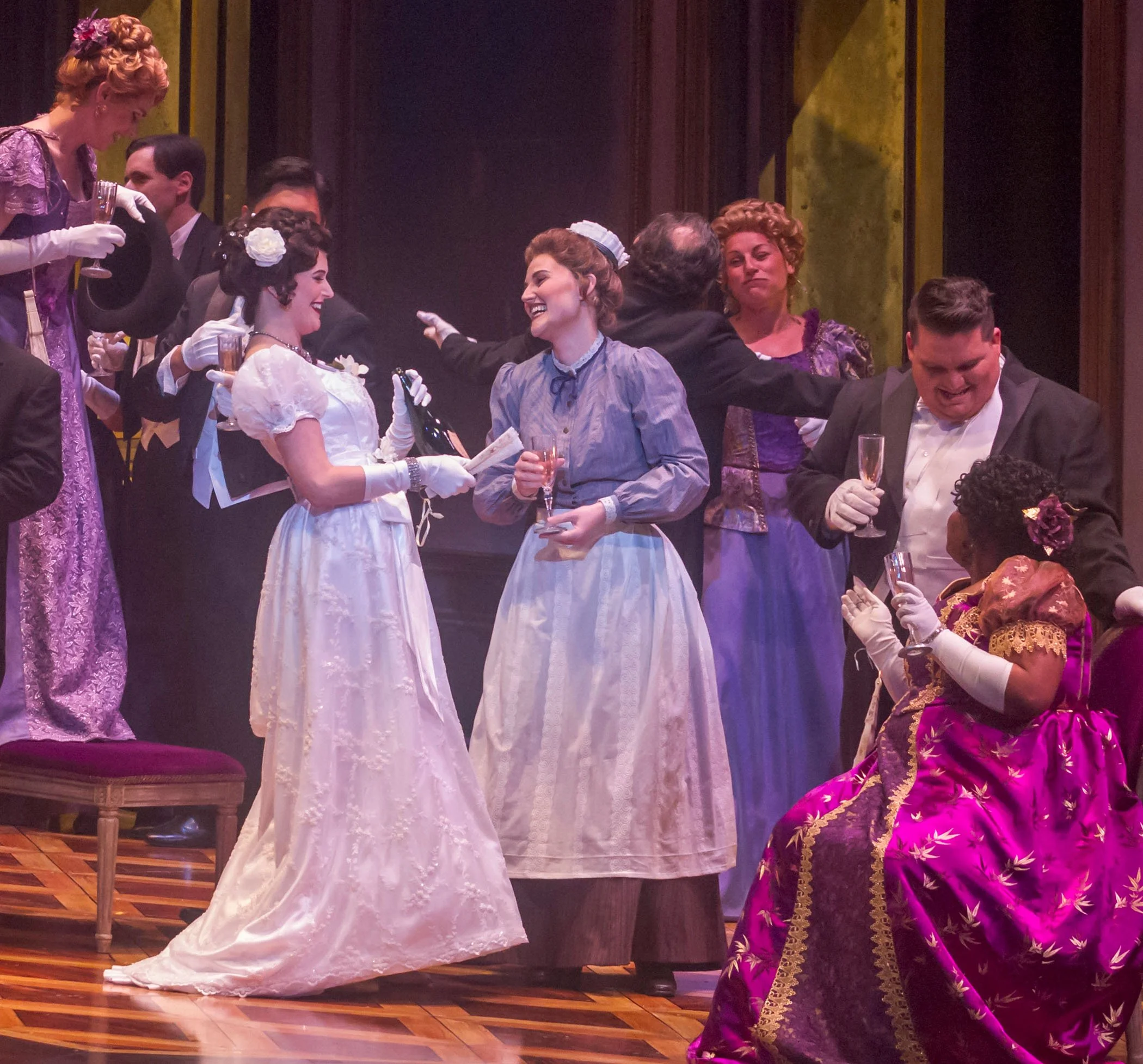 Theatrical stage scene with actors in historical costume celebrating, holding glasses of champagne, smiling and chatting at a formal gathering.