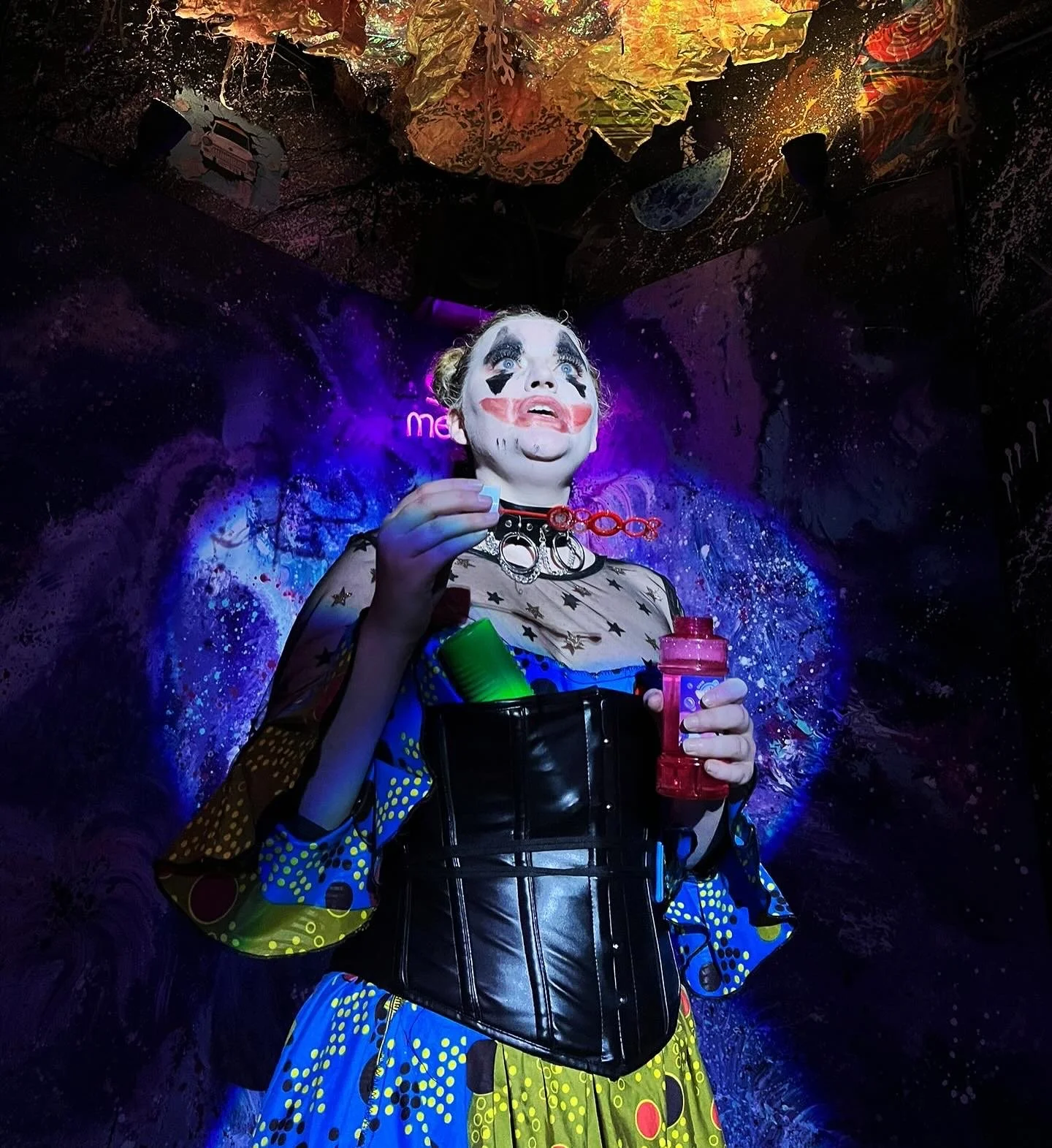 A person dressed as a clown with painted face makeup, holding a bottle of bubbles and a bubble wand, standing in front of a colorful cosmic-themed background with art and neon lighting.