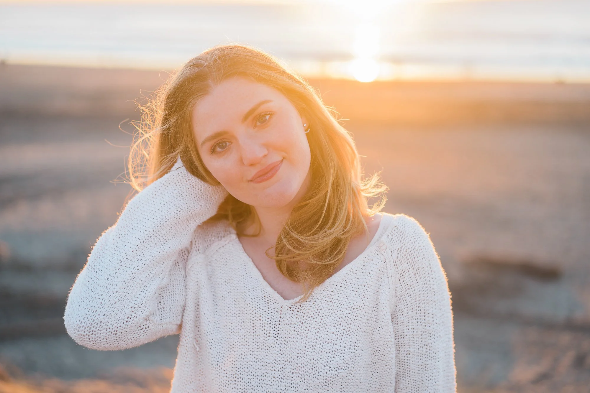 A young woman with light brown hair and fair skin wearing a white sweater, standing on a beach during sunset, with the ocean in the background and sunlight illuminating her face.