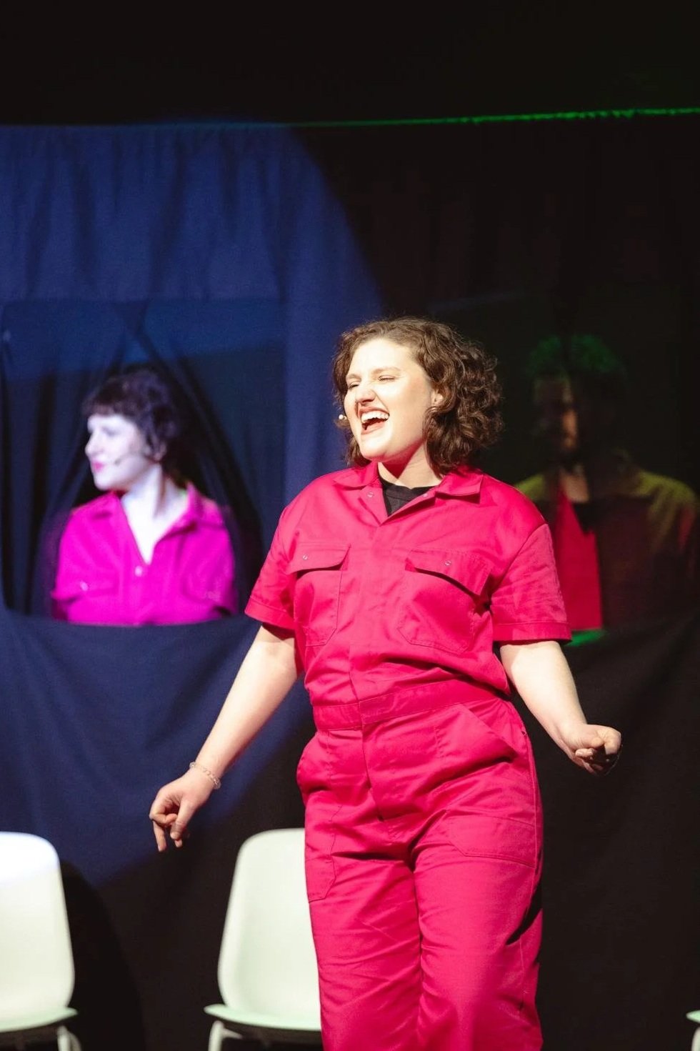 A woman in a red jumpsuit singing passionately on stage, with two reflected images behind her, one of which has short curly hair and a pink shirt, both on dark backgrounds.