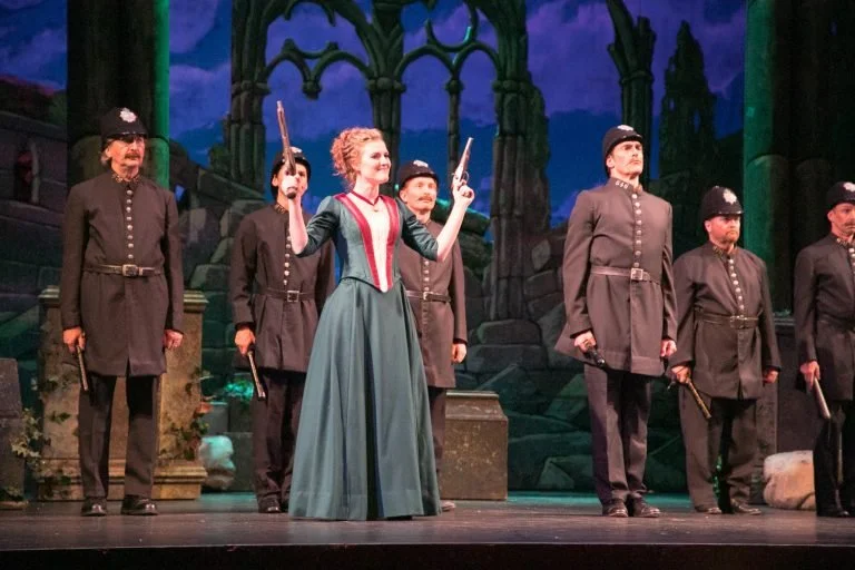 A theatrical stage scene with a woman in period costume holding two guns, surrounded by six men in uniform, against a backdrop of ruins and dark sky.