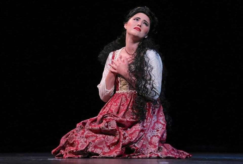 A woman dressed in a period costume, sitting on the floor against a black background, looking upwards with an emotional expression.
