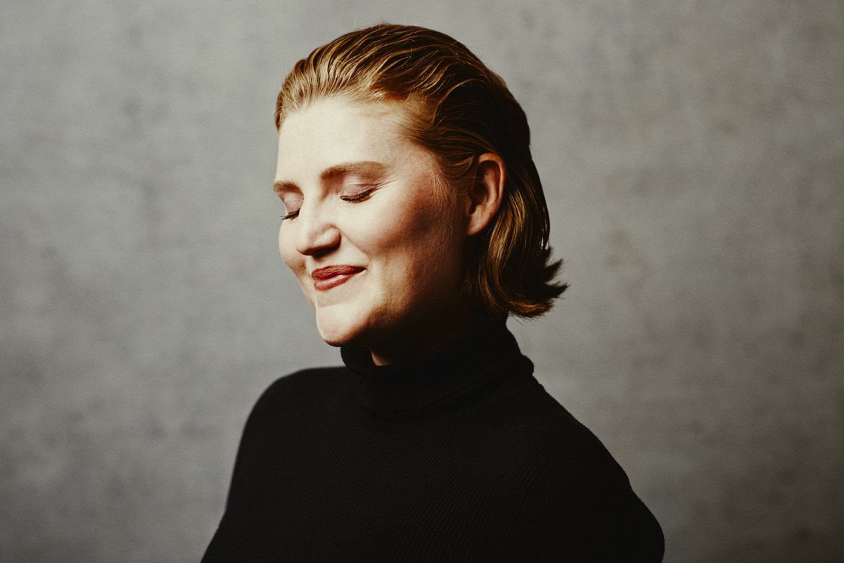 A woman with short red hair in a black turtleneck, eyes closed, smiling softly against a plain gray background.