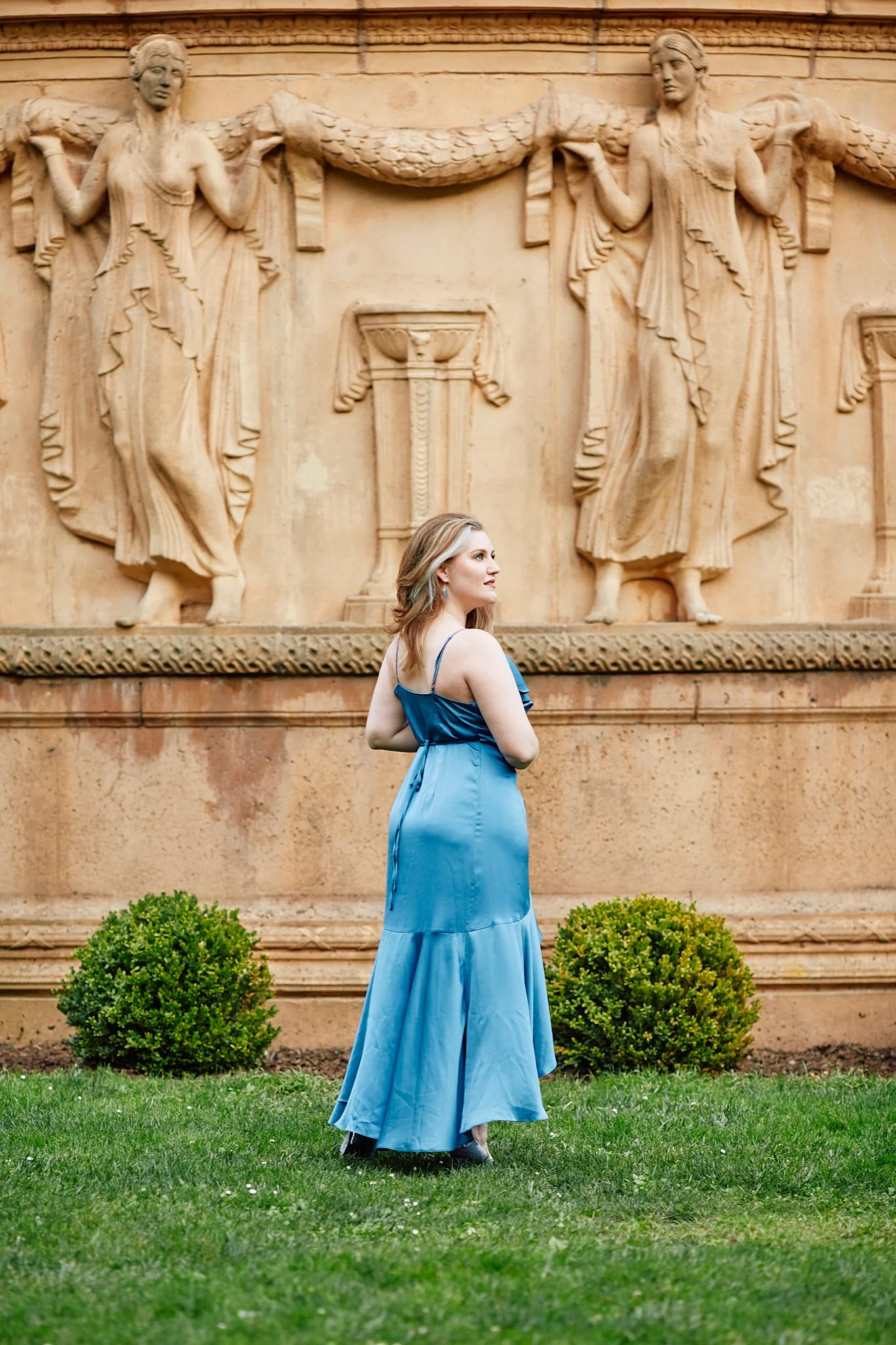 A woman in a blue dress standing on grass in front of a stone wall with classical-style sculptures of women holding a draped fabric.