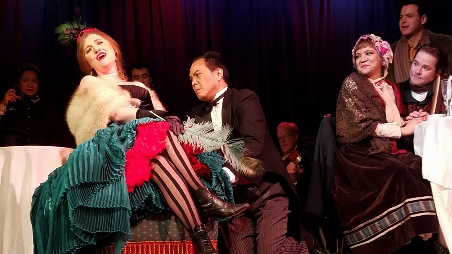 Actors on stage performing a comedic scene at a dinner theater, with one woman in a colorful, elaborate costume laughing and another man in a tuxedo listening attentively.