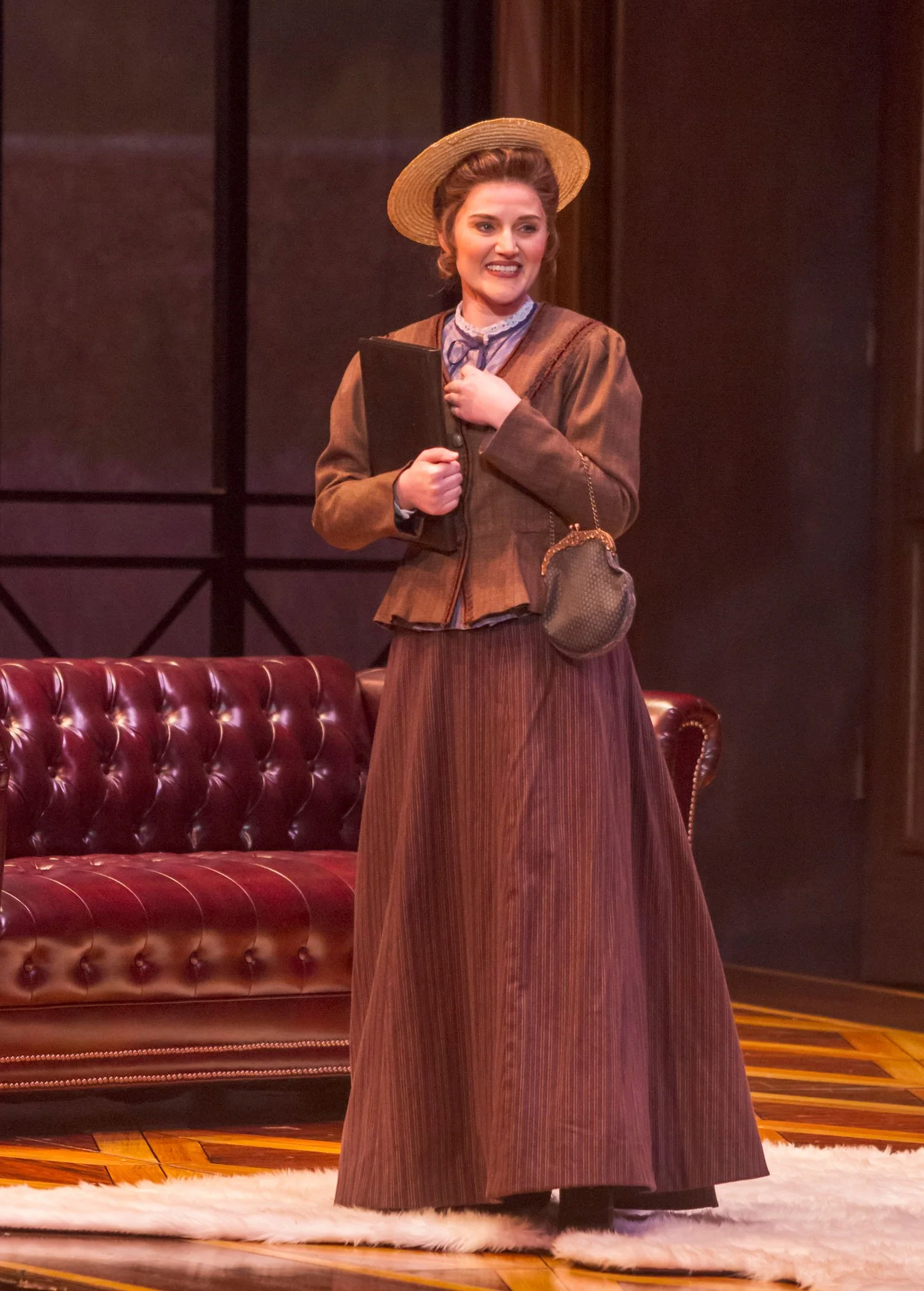 A woman dressed in vintage clothing, wearing a brown jacket and long skirt, holding a book, and standing in front of a leather sofa.