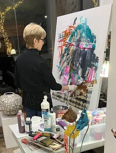 Woman with short blonde hair painting an abstract colorful artwork on a large canvas with brushes in an art studio.