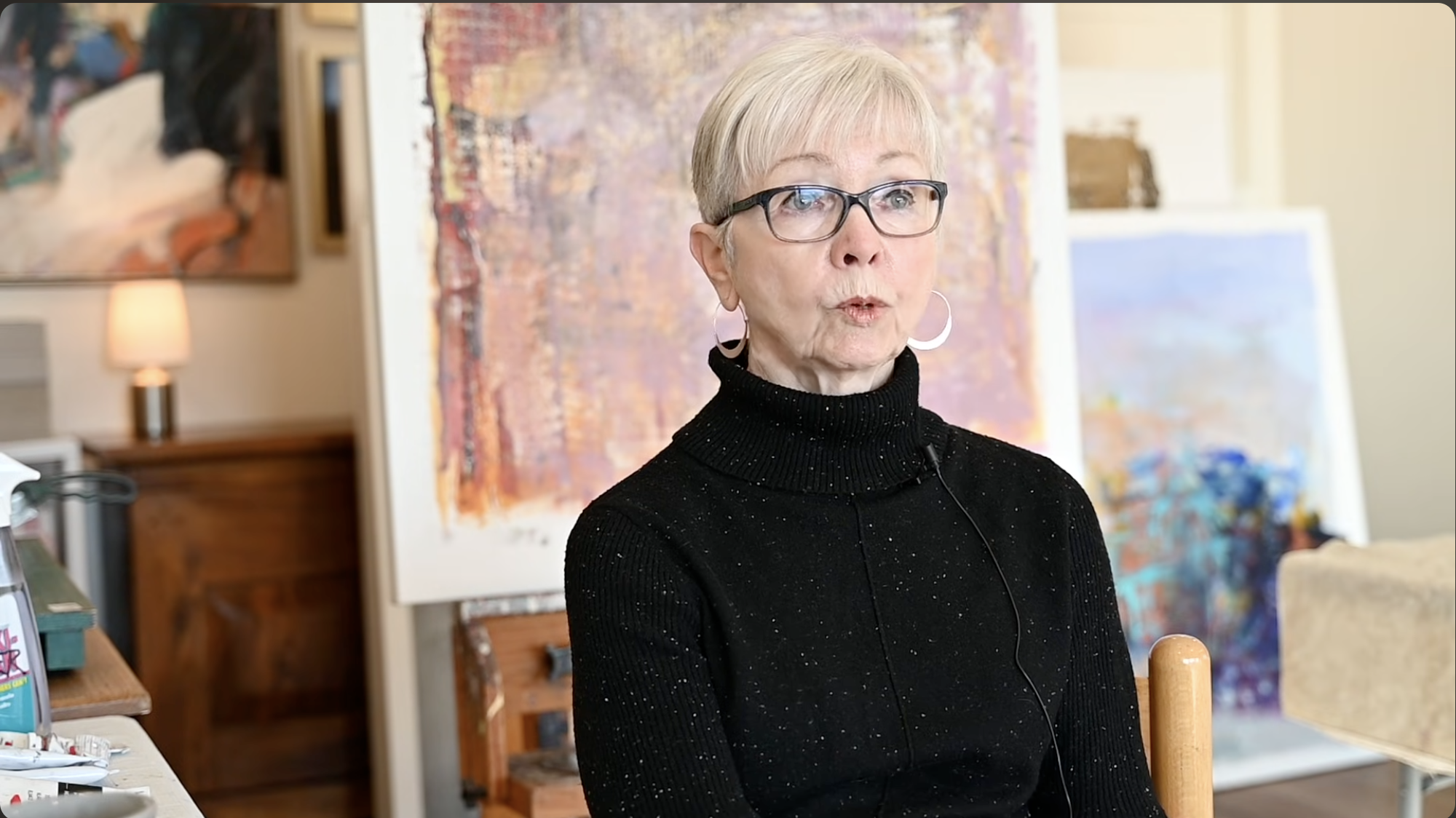 An elderly woman with short gray hair, glasses, and hoop earrings, wearing a black turtleneck sweater, seated indoors in front of colorful abstract paintings, with a lamp and furniture visible in the background.