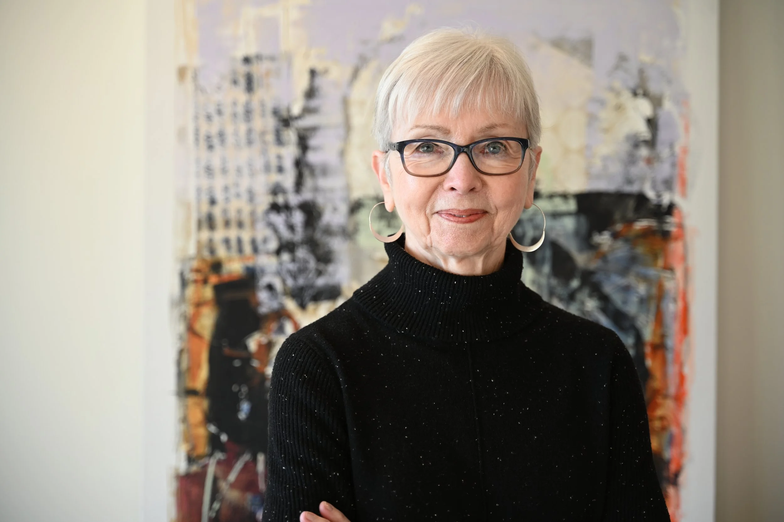 An elderly woman with short gray hair wearing glasses, a black turtleneck sweater, and hoop earrings, standing in front of an abstract art painting.