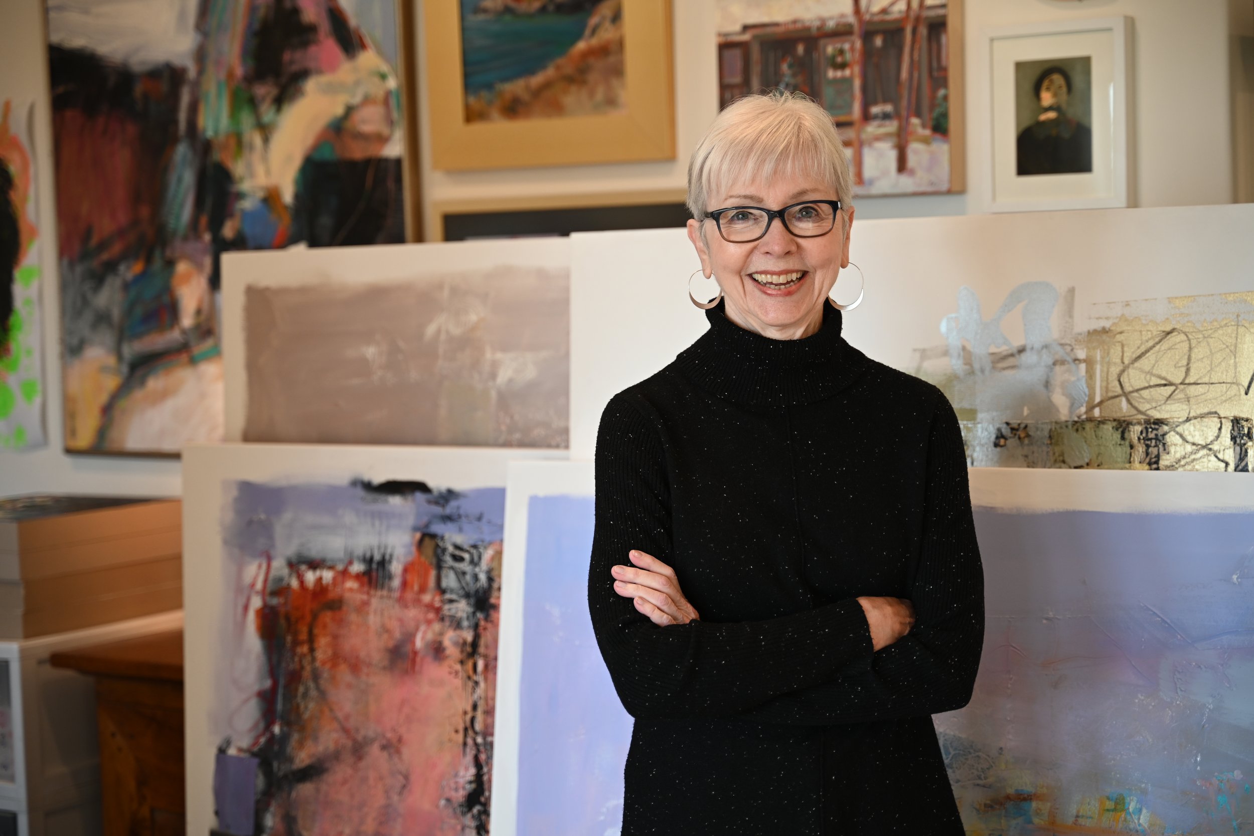 An elderly woman with short gray hair, glasses, and hoop earrings, standing in front of abstract paintings in an art gallery, smiling with her arms crossed.