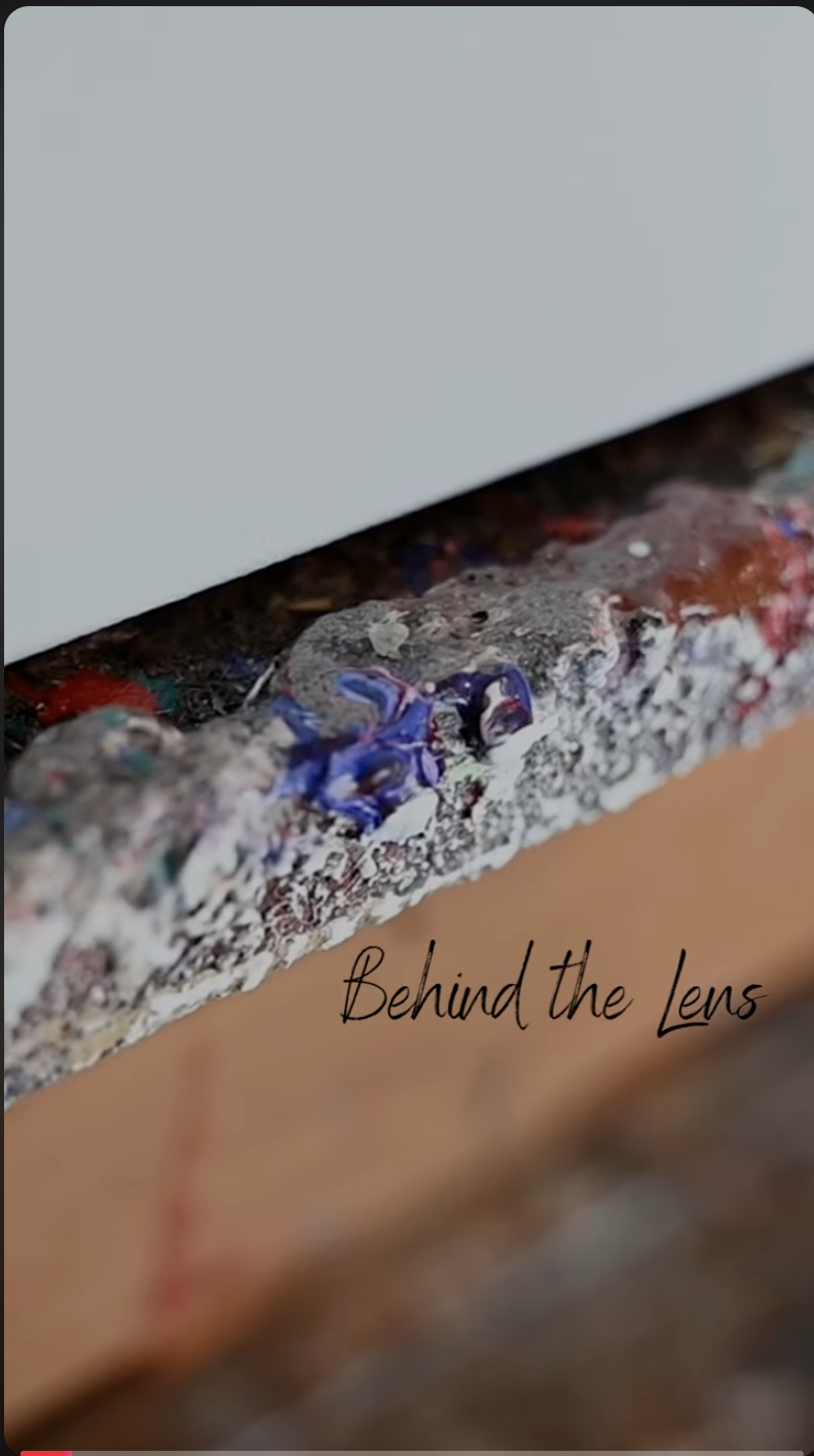 Close-up of a cluttered painting workspace with spilled paint on a textured surface and the words "Behind the Lens" written in black cursive font.