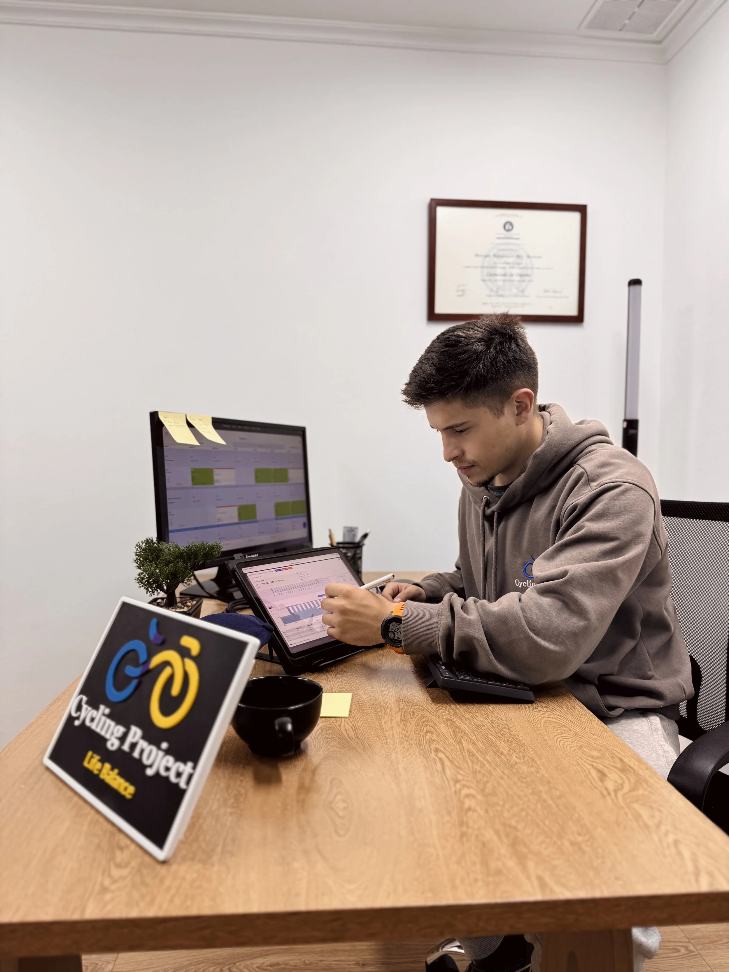 Coach sentado en una mesa de oficina, mirando una tableta, con una computadora, una maceta pequeña y un bolígrafo. En el fondo, un marco con un diploma y un cartel con un símbolo de bicicleta y el texto 'Cycling Project Uzhhorod'.