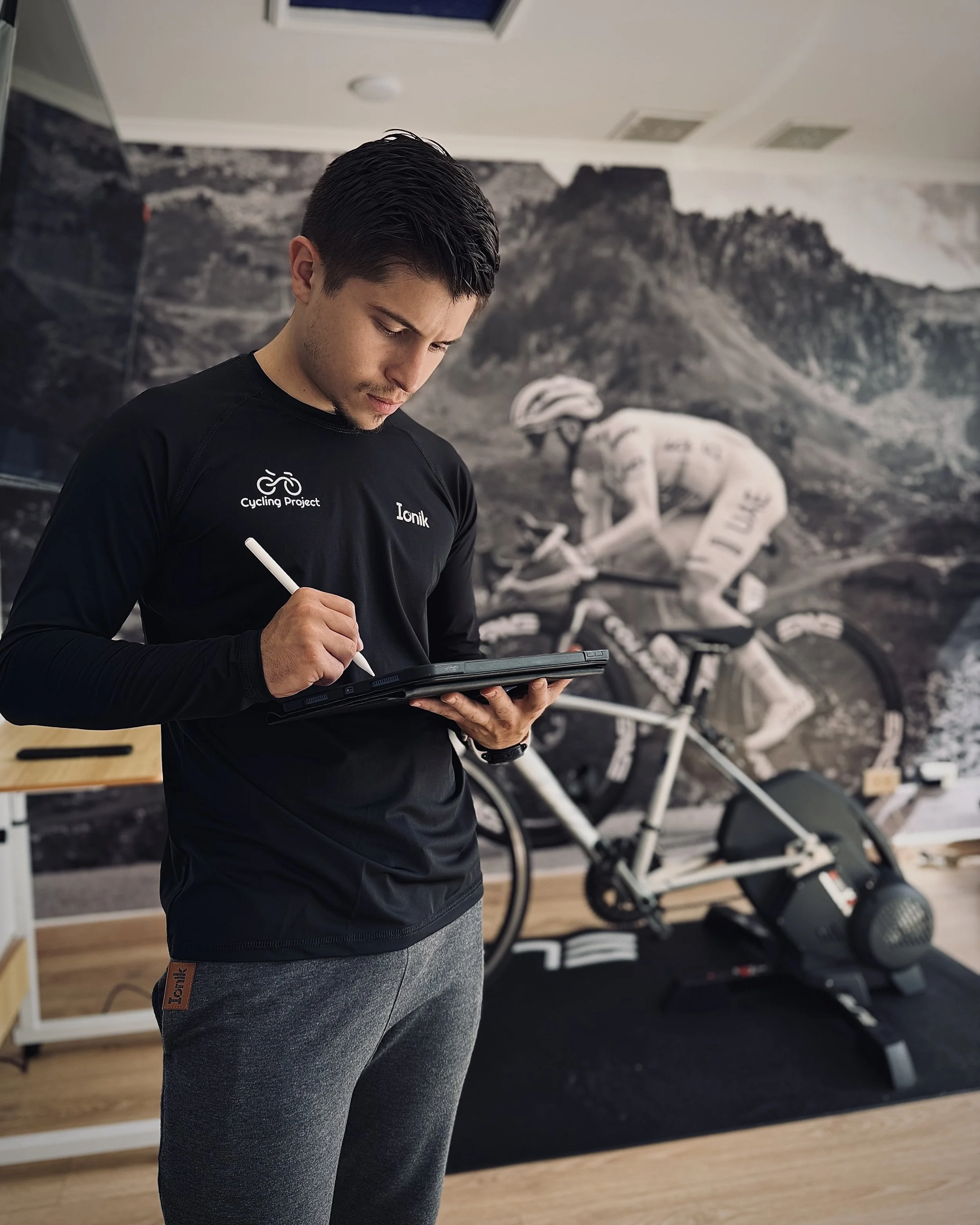 Un joven con camiseta negra que dice 'Cycling Project' y 'LoniK' en una sala de entrenamiento, escribiendo en una tableta digital, con una bicicleta en el fondo y un mural de montañas.