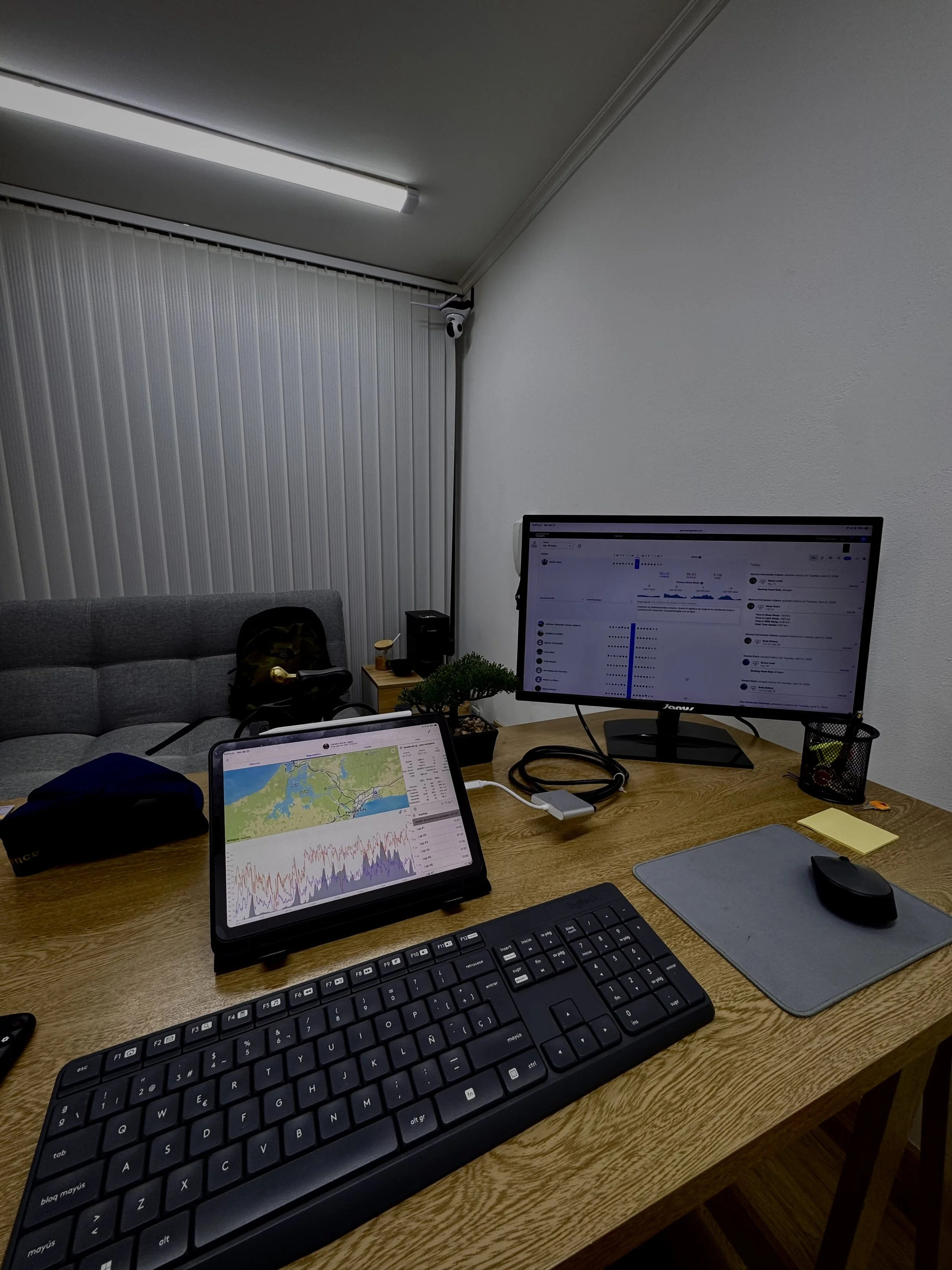 Mesa de oficina con computadora de escritorio y portátil, teclado, ratón, cuaderno, y otros objetos, en una habitación con paredes blancas y ventanas con persianas verticales.