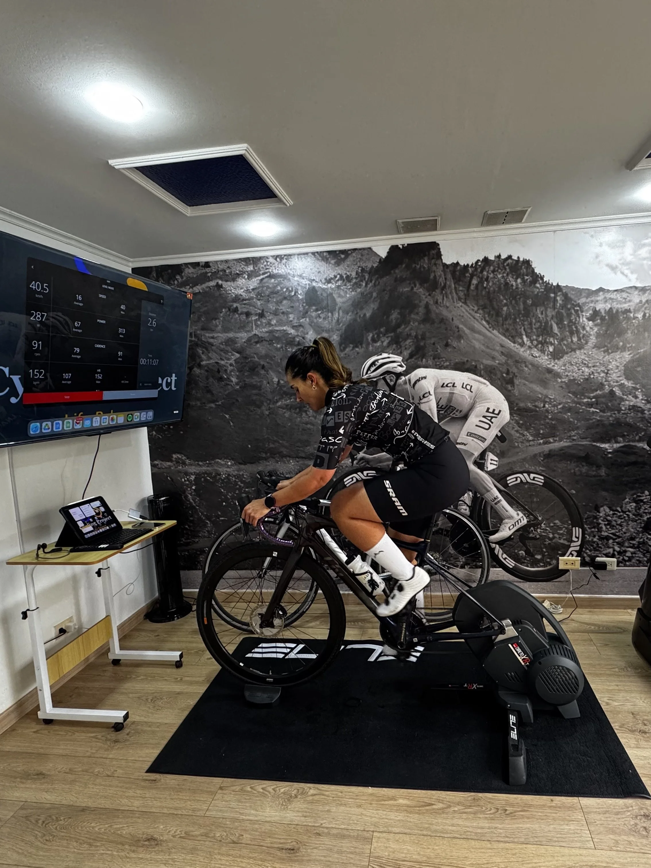 Persona haciendo ejercicio en una bicicleta de spinning en una sala con mural de montañas y una pantalla con datos de entrenamiento.