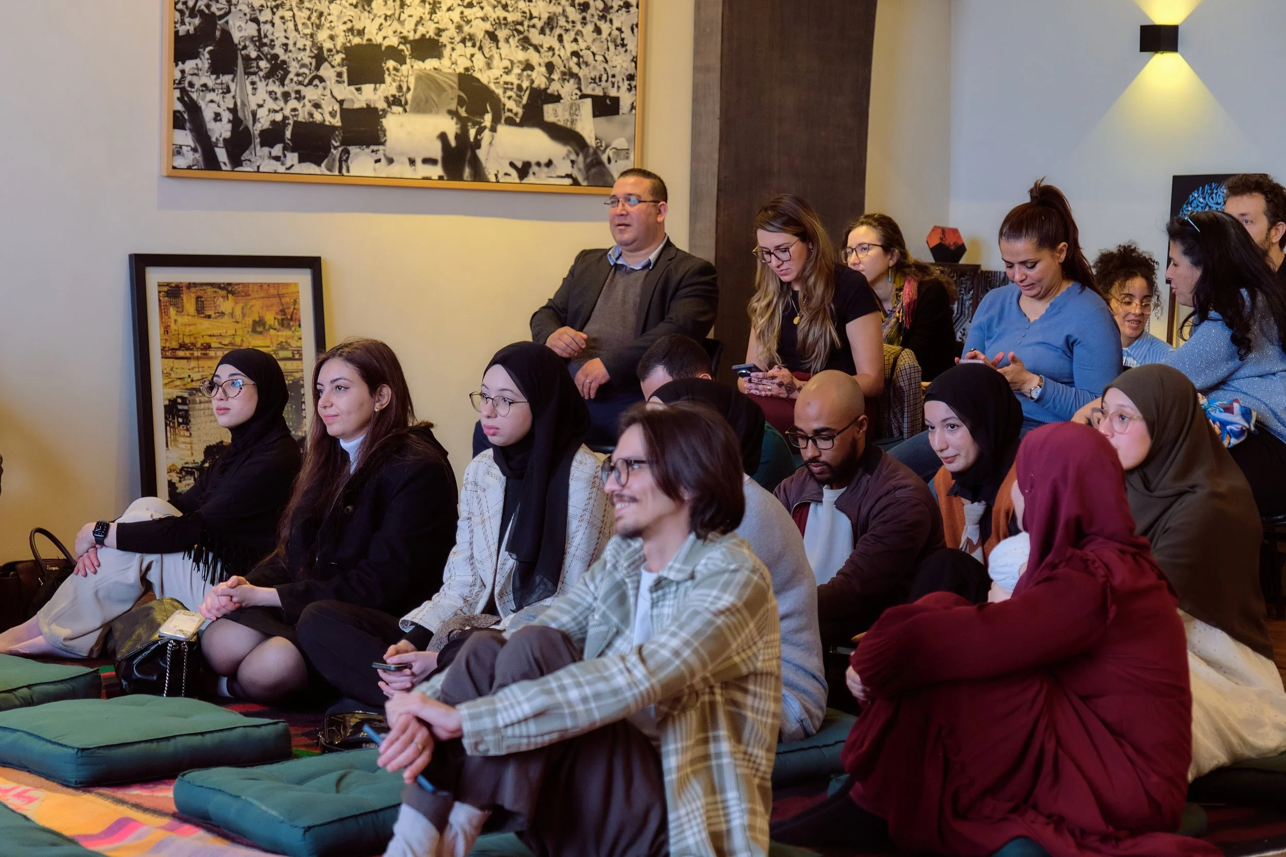 Groupe de personnes assises et debout lors d'un événement ou d'une conférence en intérieur, certains utilisant leur téléphone.