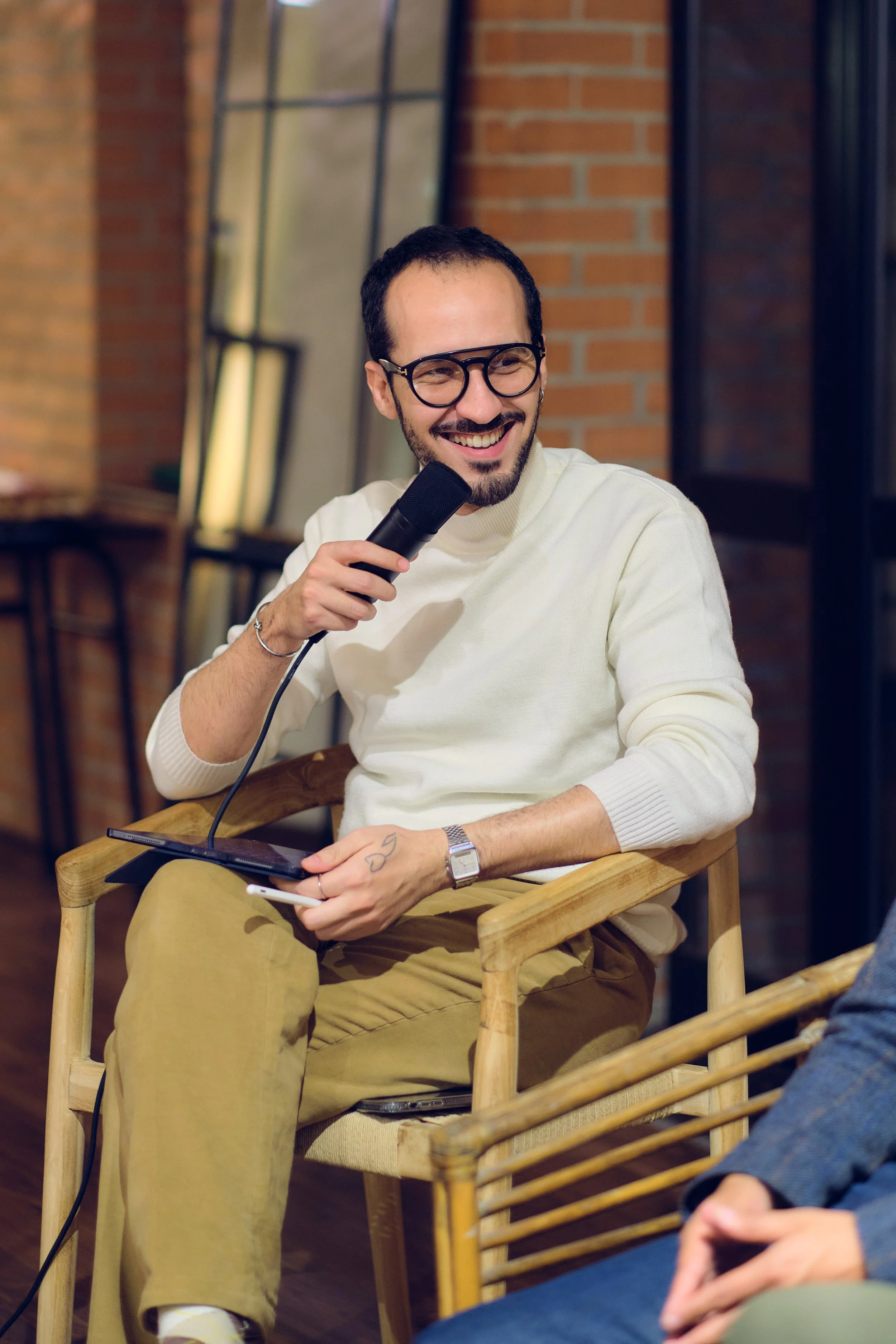 Un jeune homme souriant, portant des lunettes noires et une chemise blanche, tient un microphone et semble parler lors d'une interview ou d'une présentation. Il est assis dans un fauteuil en bois dans un intérieur avec un mur en briques.
