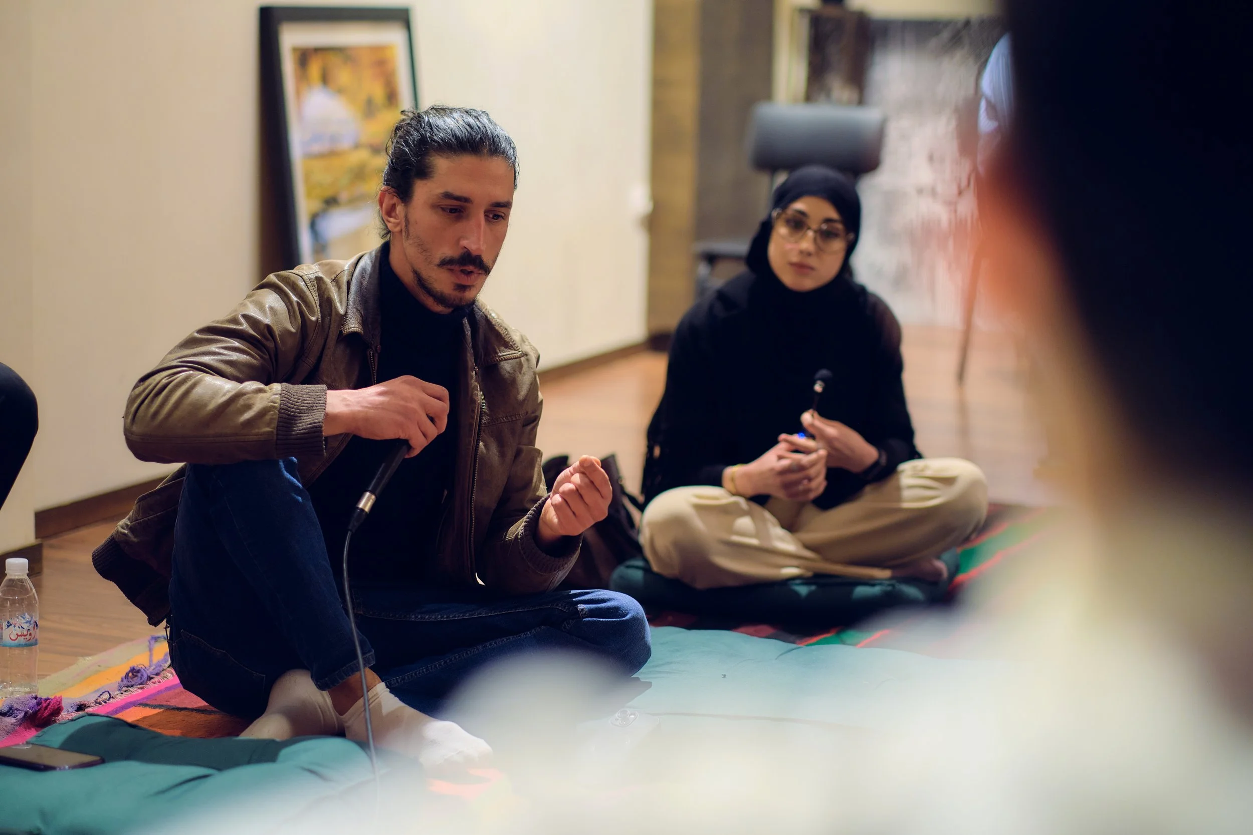 Deux personnes, un homme et une femme, assissent sur un tapis au sol dans une pièce, participant à une discussion ou un atelier.