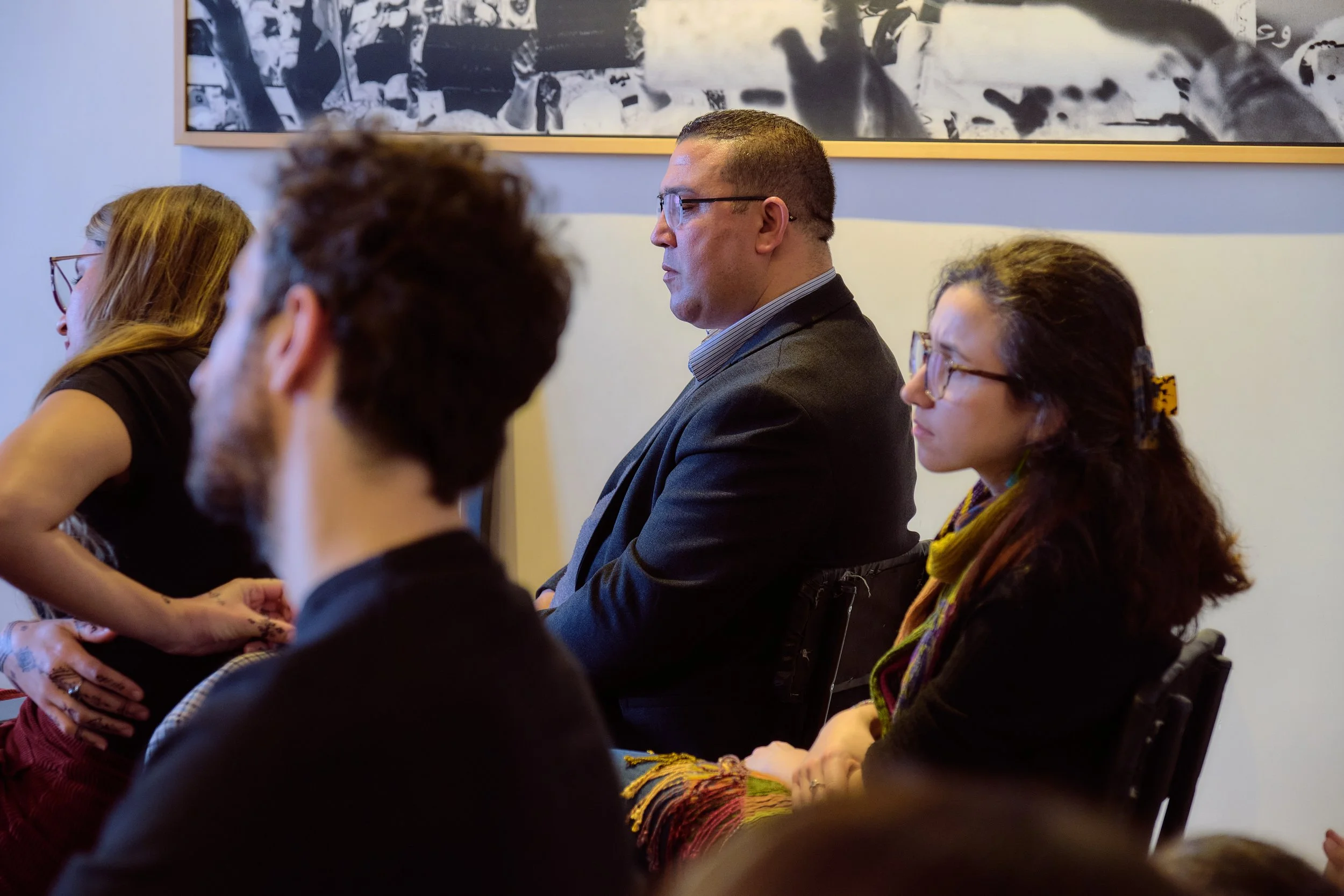 Groupe de personnes assises lors d'une réunion ou conférence, dont un homme en costume et plusieurs femmes portant des lunettes et des écharpes, dans une salle avec une œuvre d'art en noir et blanc en arrière-plan.