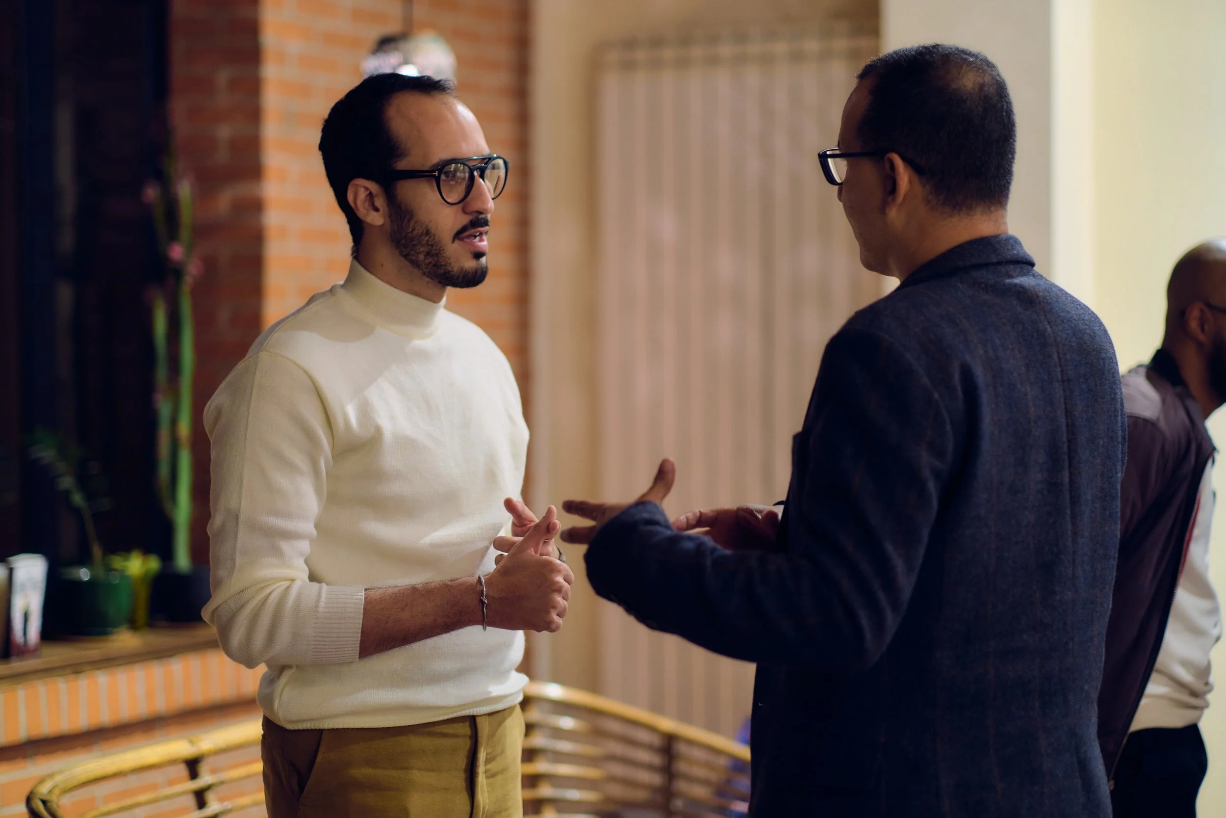 Deux hommes discutent dans un intérieur, l'un en pull blanc et l'autre en veste foncée, avec un fond en brique.