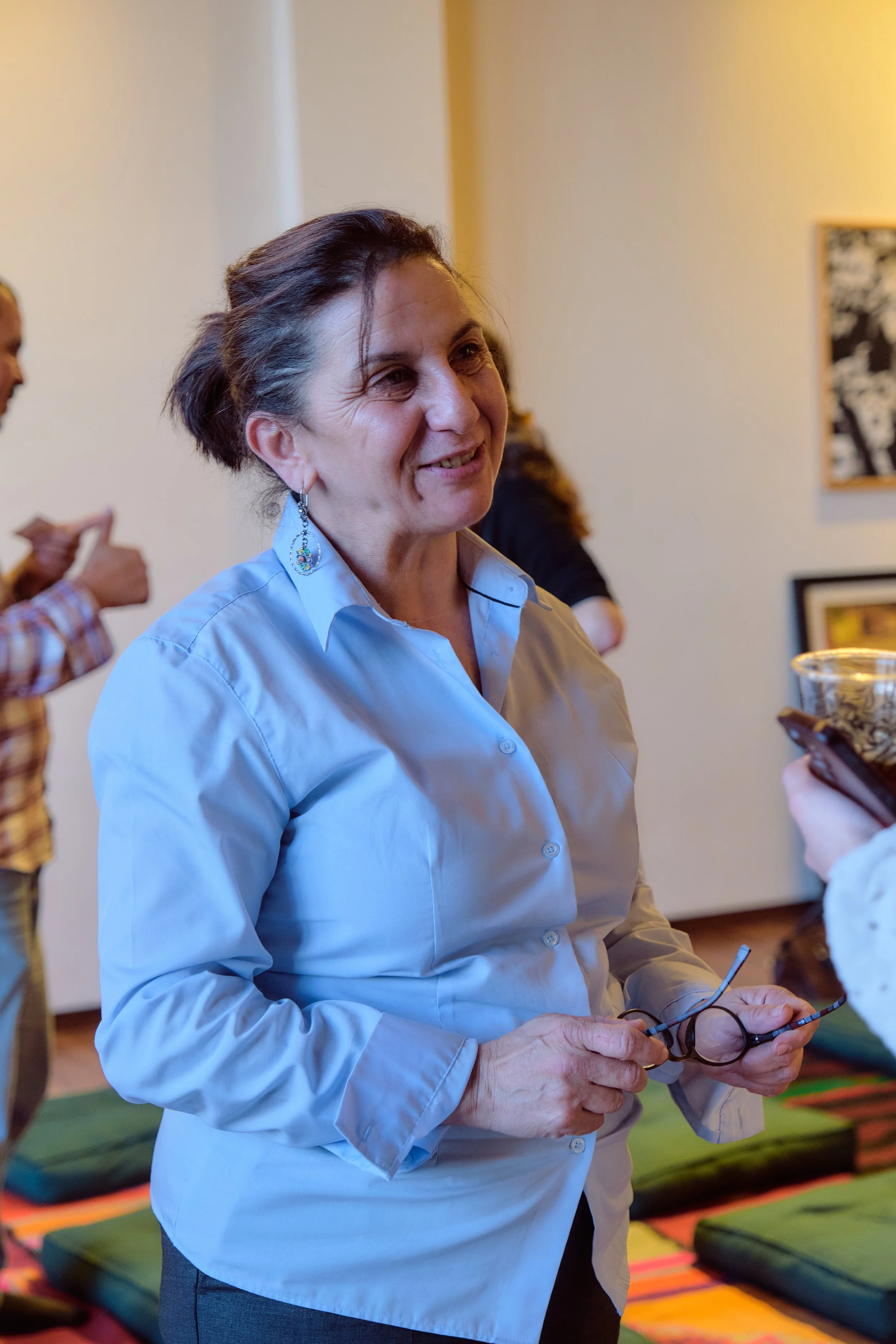 Une femme souriante avec des lunettes et des boucles d'oreilles en émail, tenant ses lunettes, lors d'une conversation en intérieur, avec plusieurs personnes en arrière-plan.