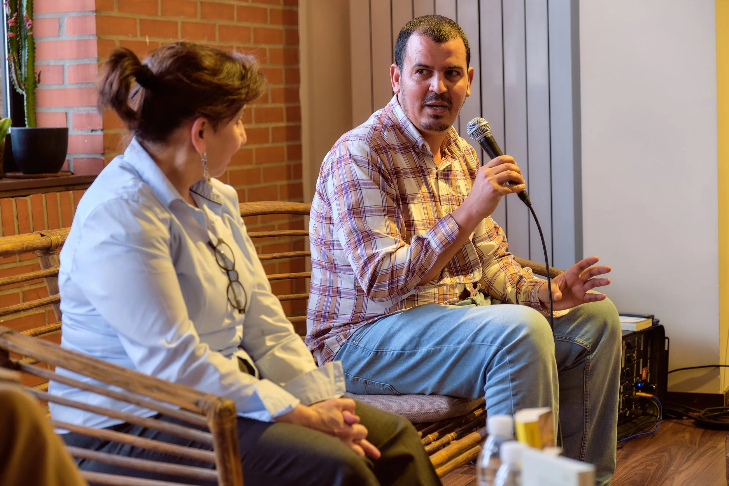 Un homme parle dans un micro lors d'une discussion, assis à côté d'une femme qui l'écoute, dans une pièce avec des murs en briques rouges.