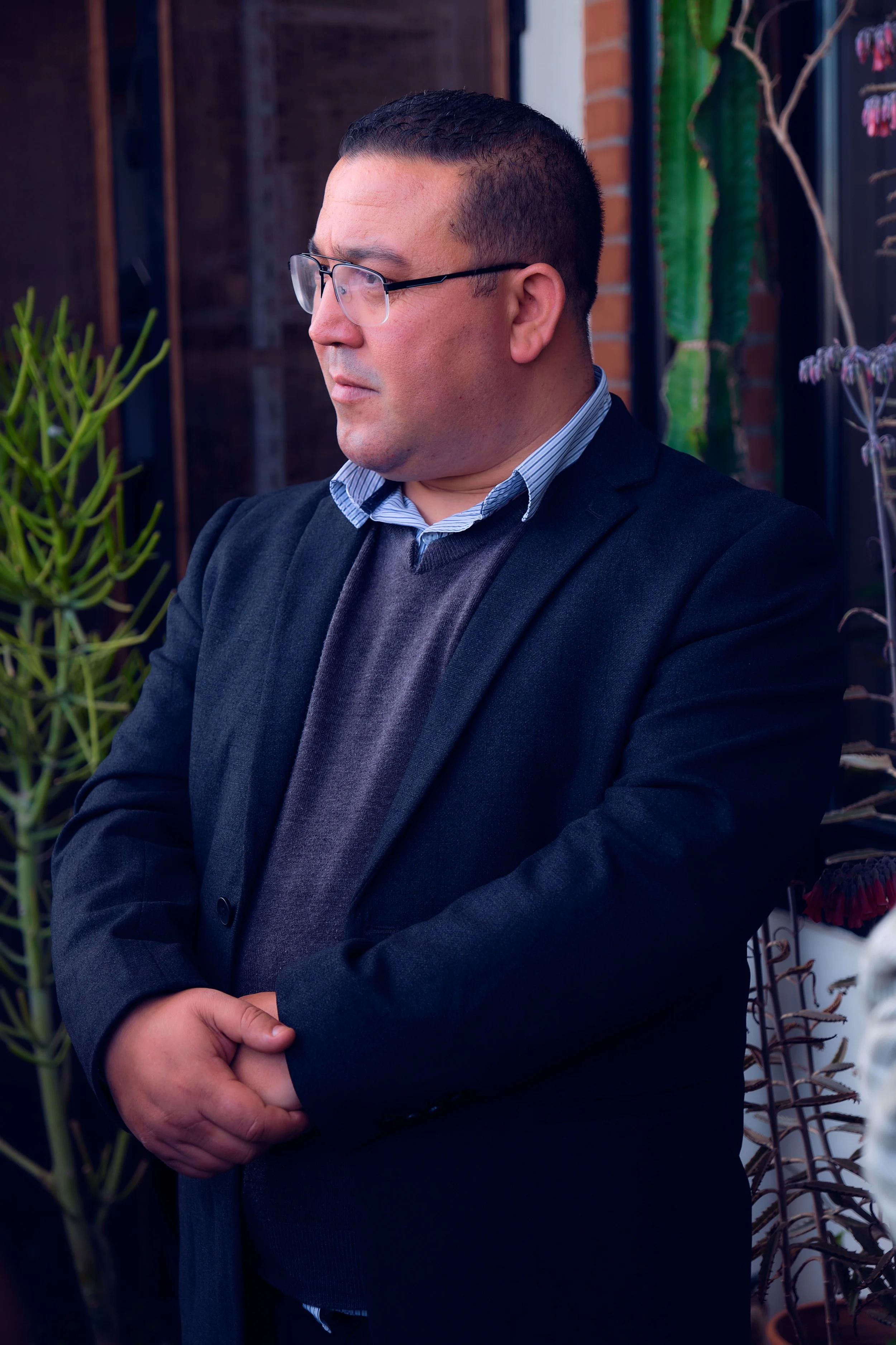 Homme portant un costume noir et une chemise à rayures, portant des lunettes, debout avec les bras croisés devant un mur en briques, avec des plantes en arrière-plan.