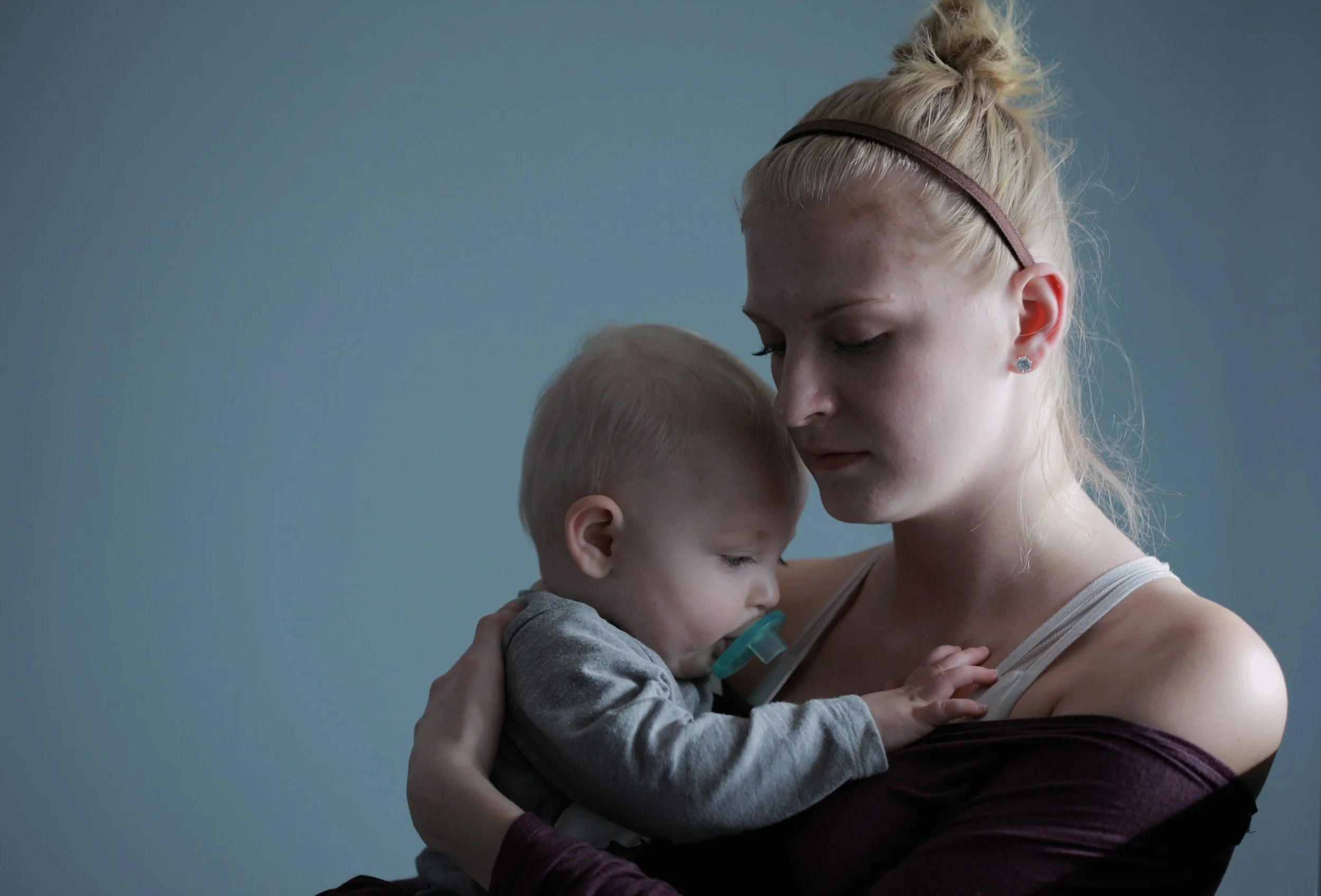 A woman with a bun hairstyle and wearing earrings is holding a young child with a pacifier, against a plain background. They are in an intimate, comforting pose, with their foreheads touching.