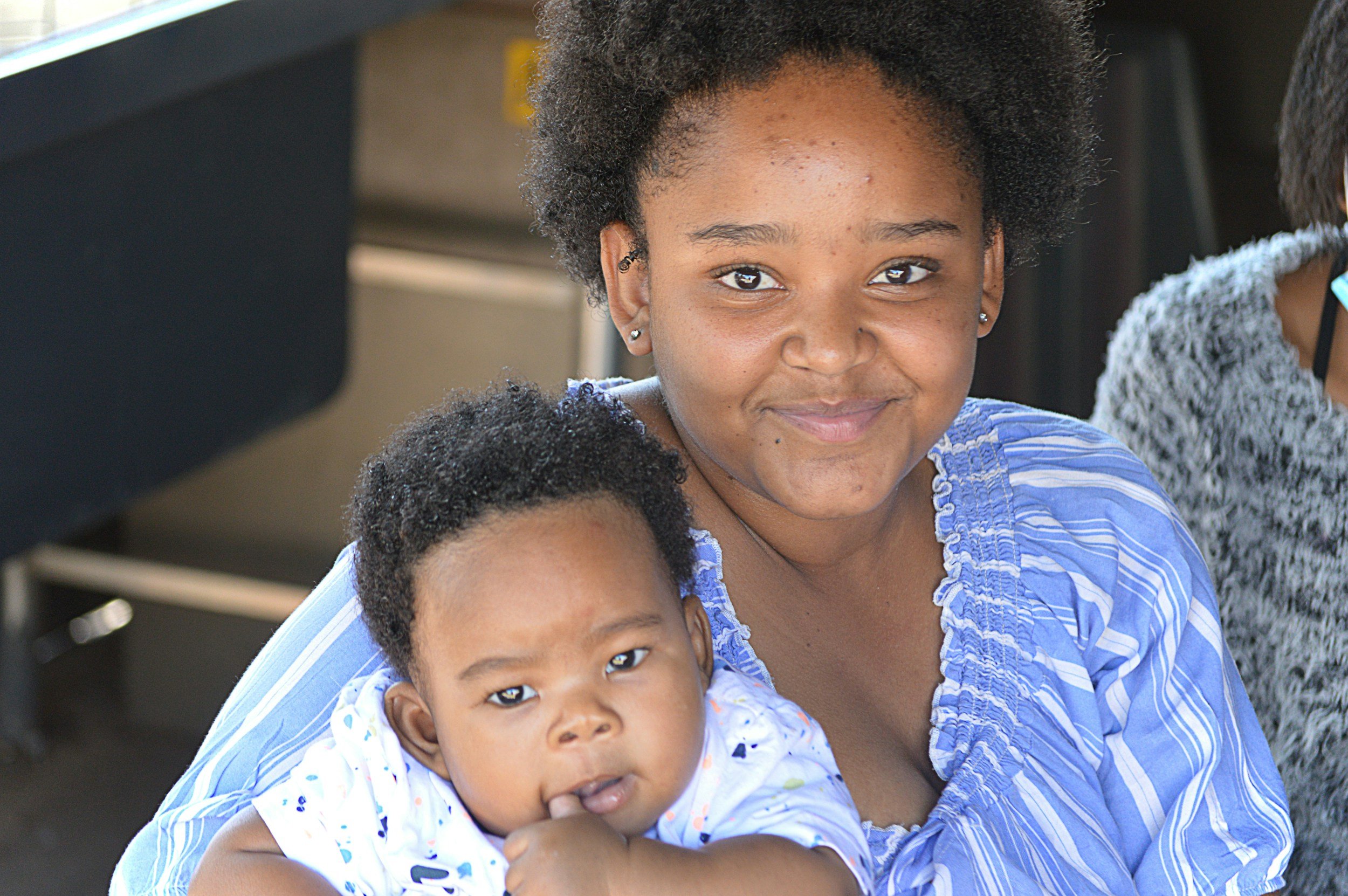 A smiling woman holding a young child in her arms, both with curly hair, sitting outdoors.