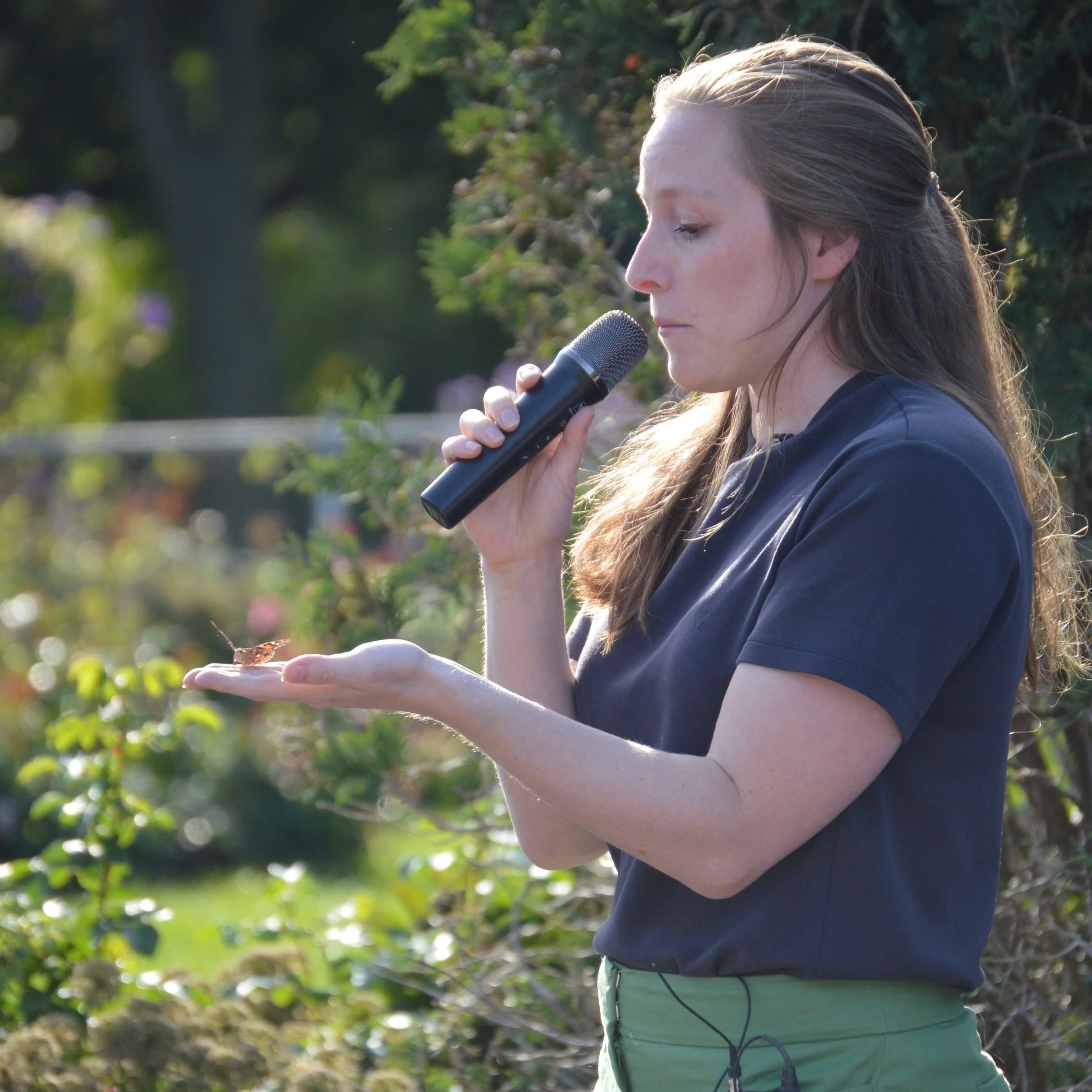 A woman with long brown hair holding a butterfly on her hand while speaking into a microphone outdoors in a garden with greenery.