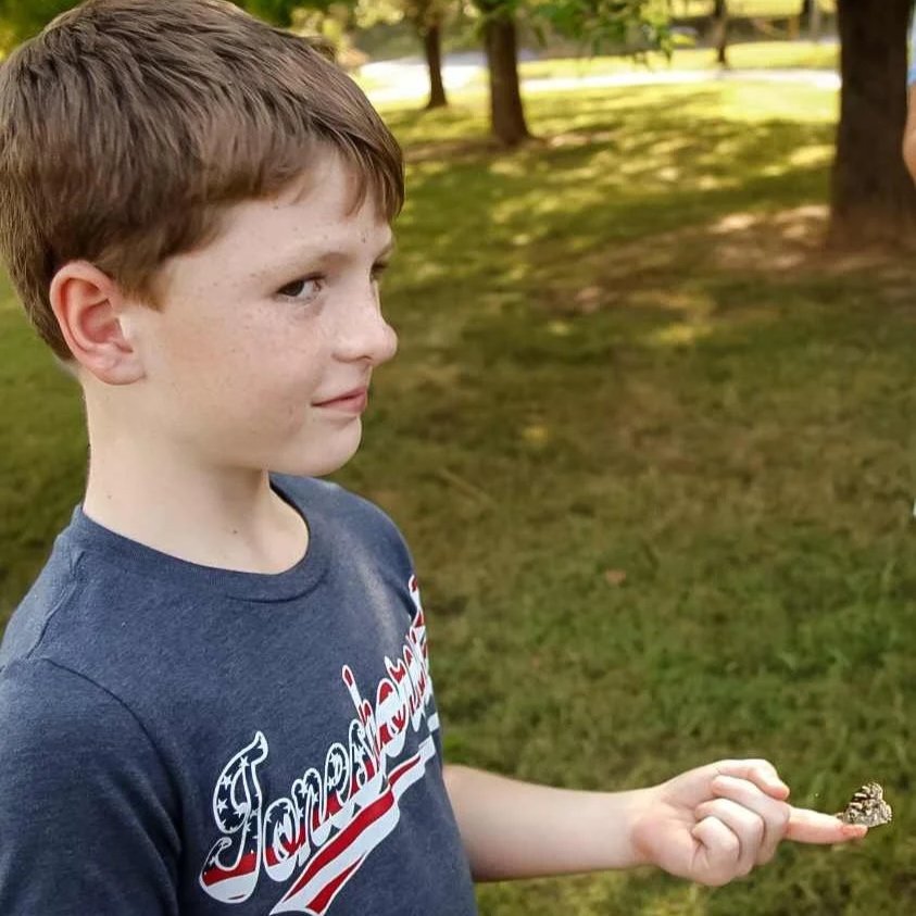 A young boy with red hair and freckles standing outdoors on grass, pointing at a small insect on his finger.