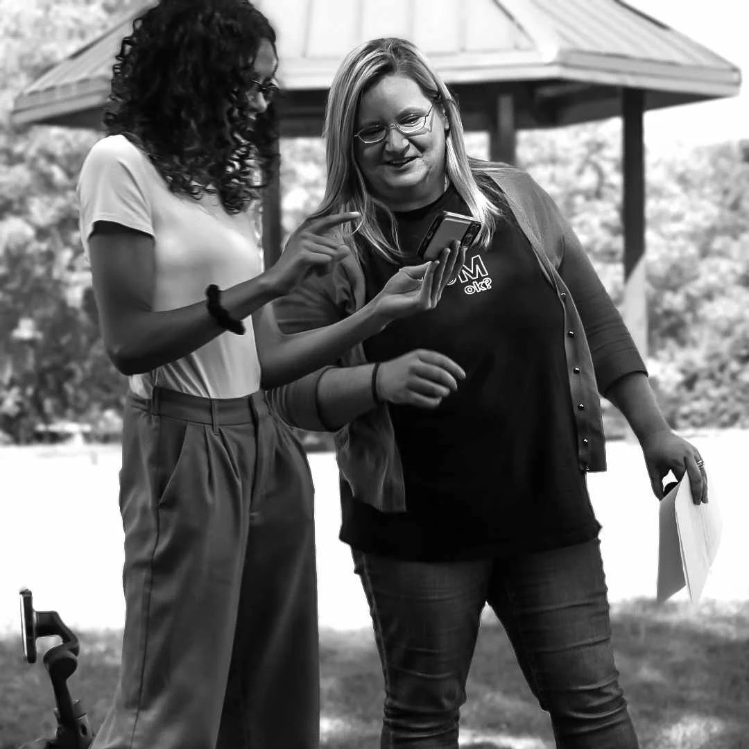 Two women standing outdoors looking at a smartphone together, with a pavilion in the background.