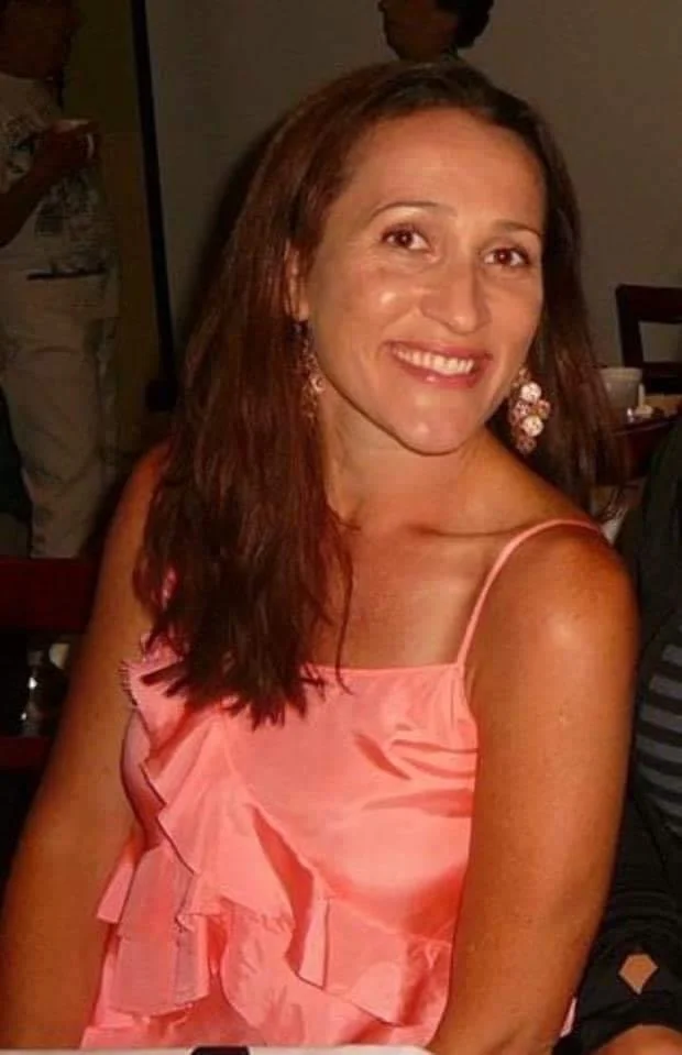 A woman with long brown hair smiling at the camera, wearing a pink ruffled spaghetti strap dress, in a restaurant or party setting.