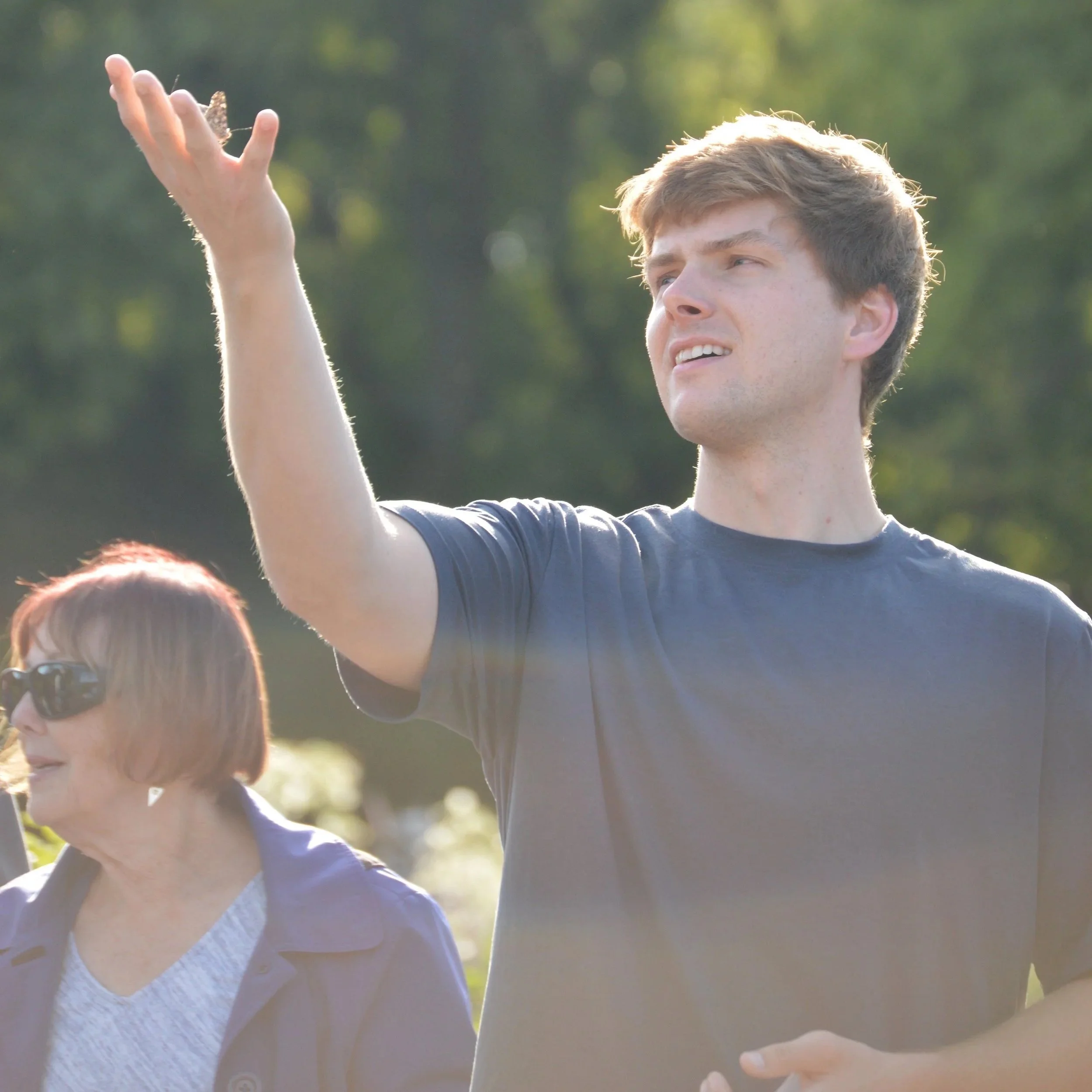 A young man in a dark T-shirt gesturing with his hand outdoors on a sunny day, with an older woman with glasses nearby.