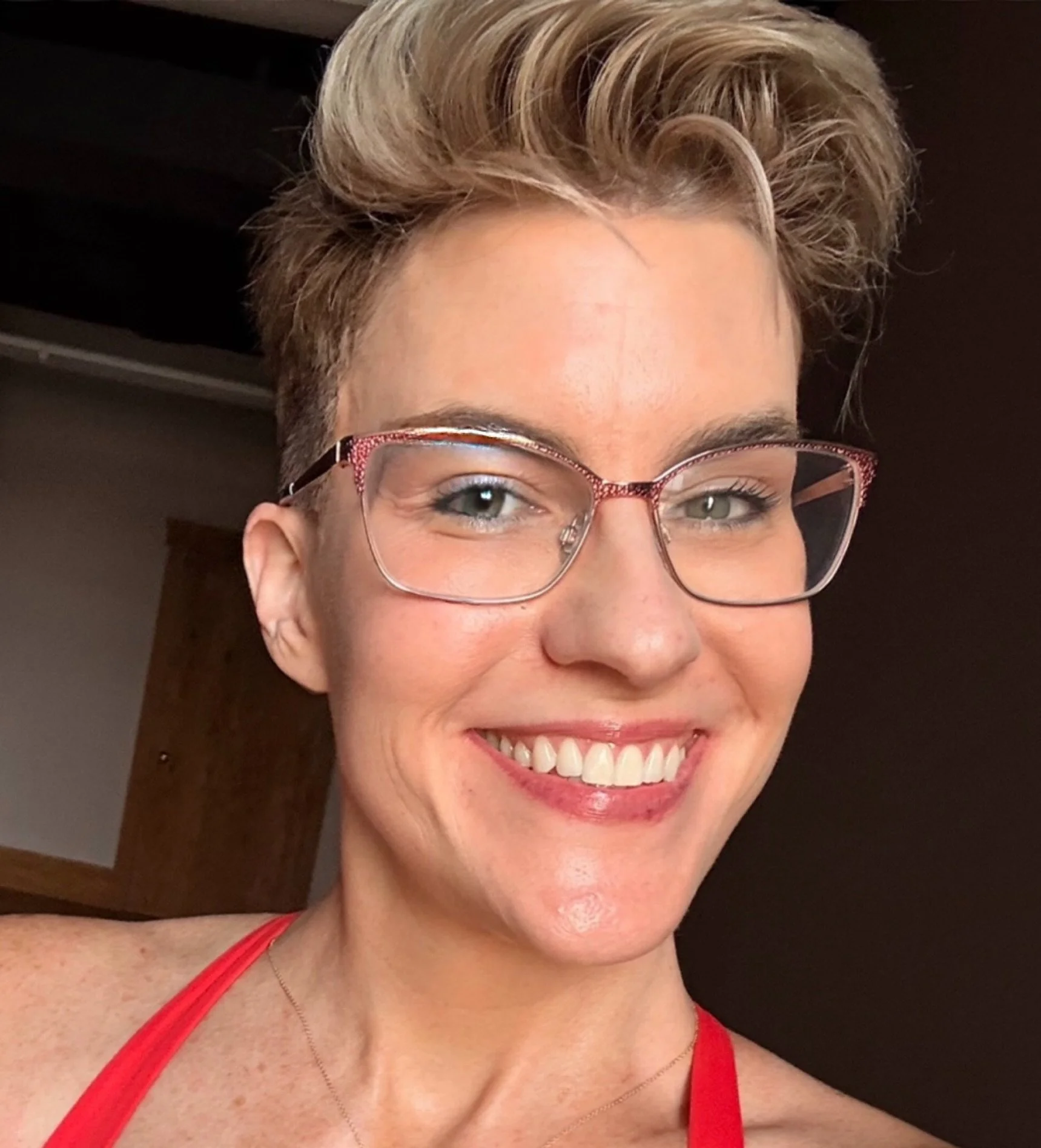 Close-up of a smiling woman wearing red top and glasses, showing her teeth and blue eyes, with short blonde hair styled in a pompadour.