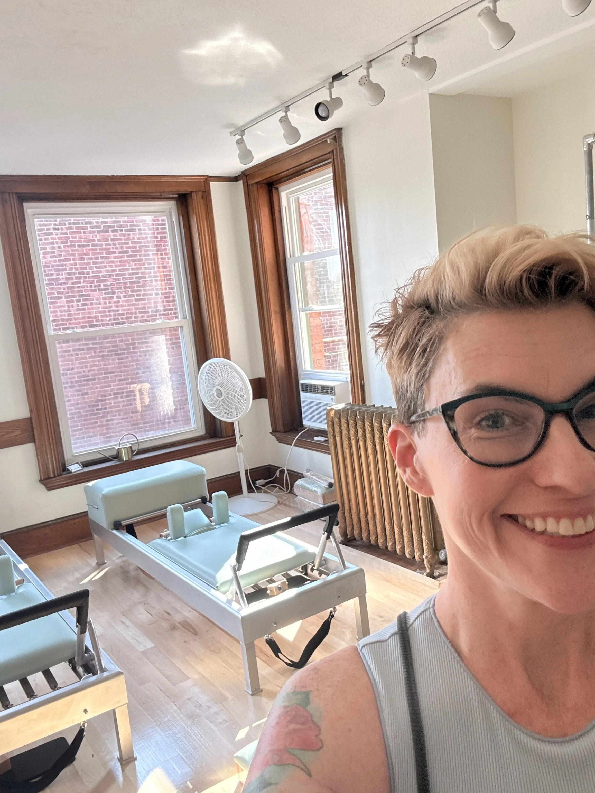 A woman smiling in a Classical Pilates Studio with light green reformers, large windows with wooden frames, and a brick building outside.