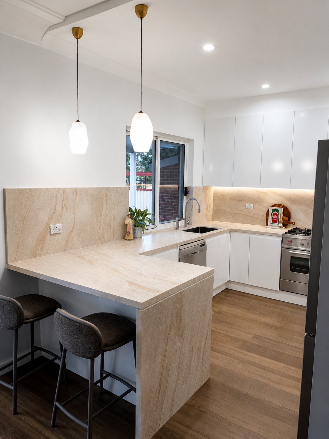 Modern kitchen with white cabinets, beige marble countertops, and wood flooring. Two pendant lights hang over a breakfast bar with two black stools. There are plants, a decorative figure, and a fire extinguisher on the counter. A window provides a vi