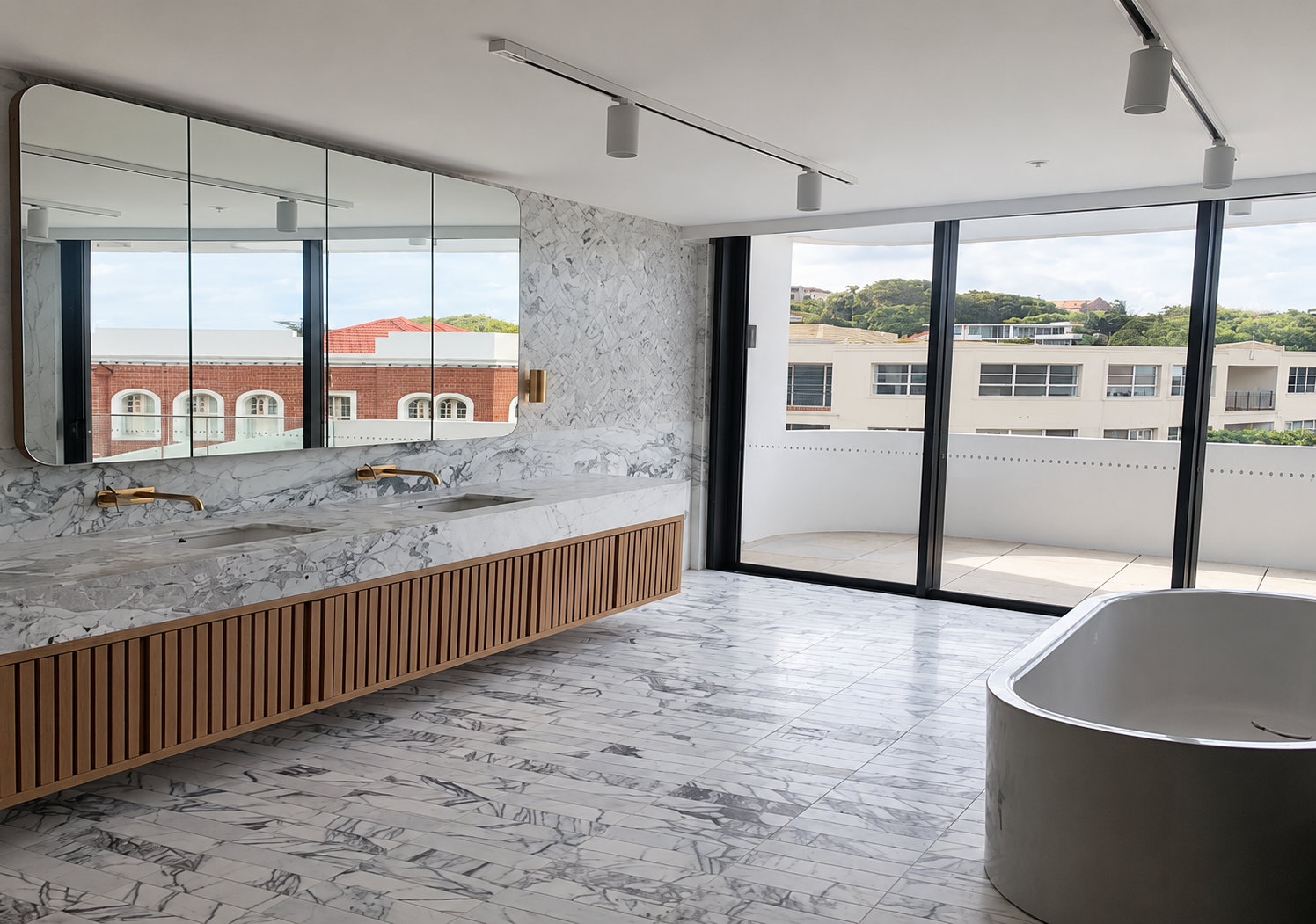 Modern bathroom with marble floors and walls, a large mirror with three sections, a marble vanity with two sinks and gold fixtures, a wooden slat cabinet below the vanity, a bathtub with marble exterior, and large sliding glass doors leading to a balcony with city and hillside views.
