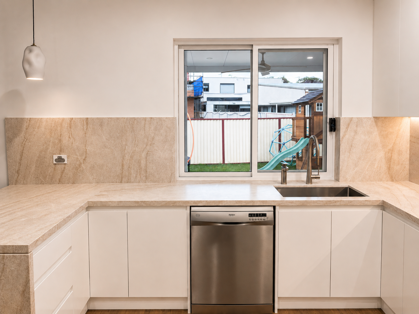 Modern kitchen with beige marble countertops, white cabinets, stainless steel dishwasher, and a large window overlooking a backyard with a slide, playhouse, and neighboring houses.