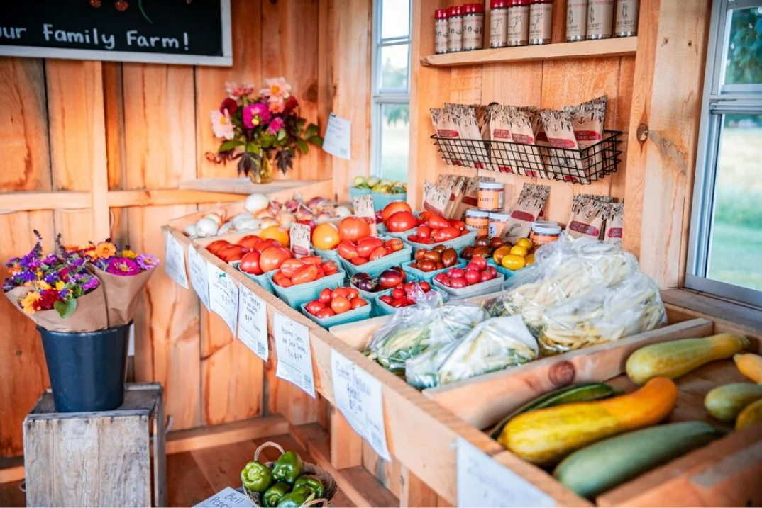 Fresh vegetables and flowers at a farm stand, including tomatoes, zucchinis, and bell peppers, with a wooden interior.