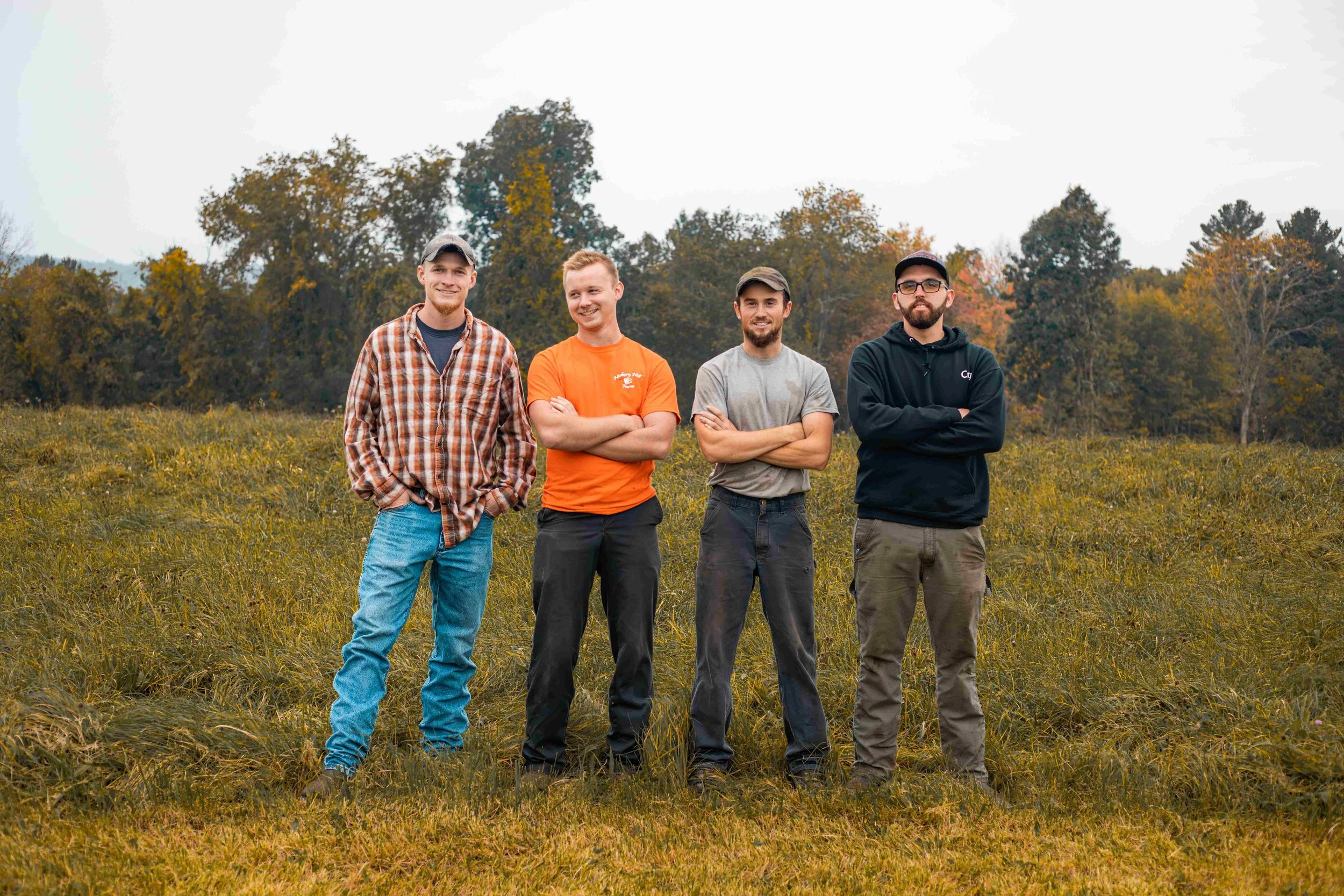 Four young men standing outdoors in a hay field with trees in the background, dressed casually and smiling.