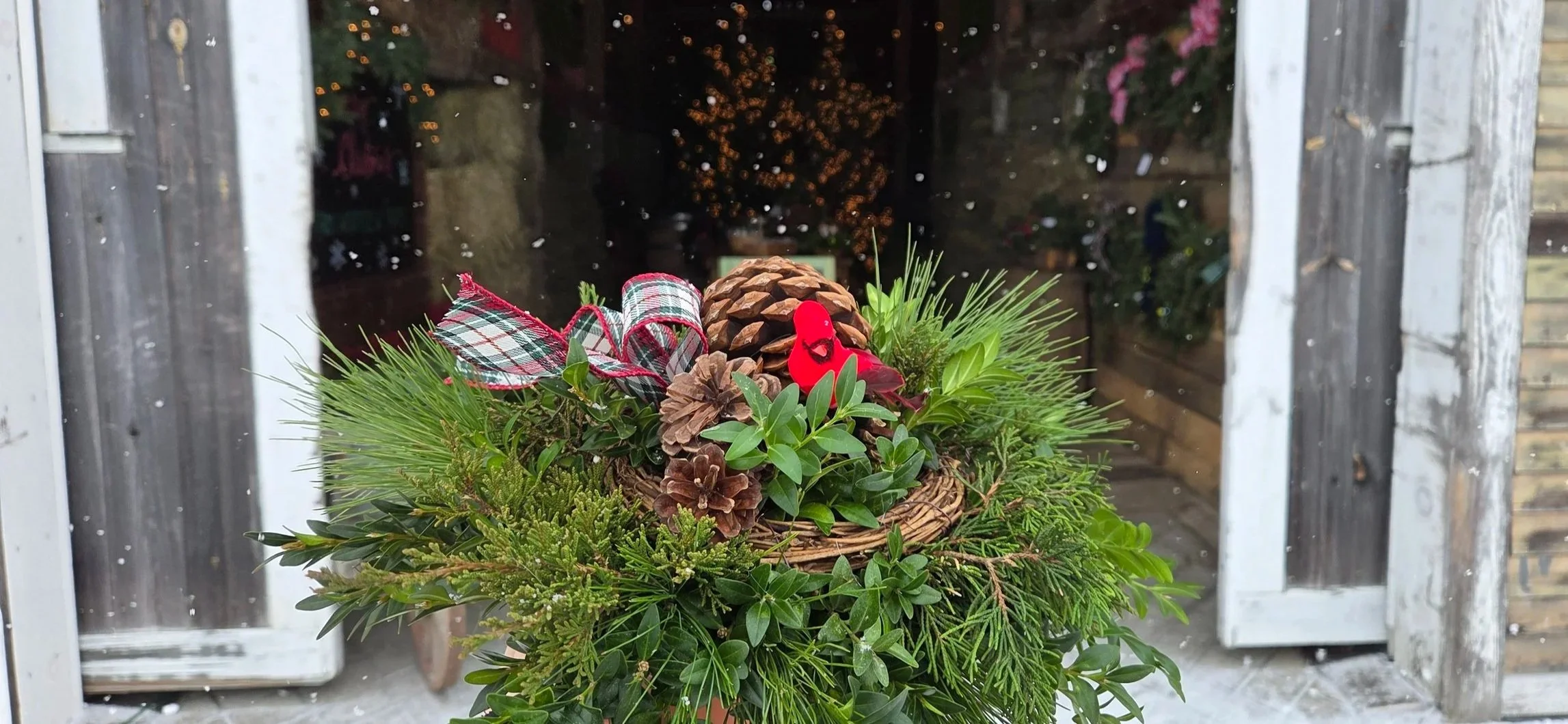 A festive holiday arrangement with pine branches, pinecones, red and green ribbon, and small red decorations, placed in front of a window with snow visible outside.