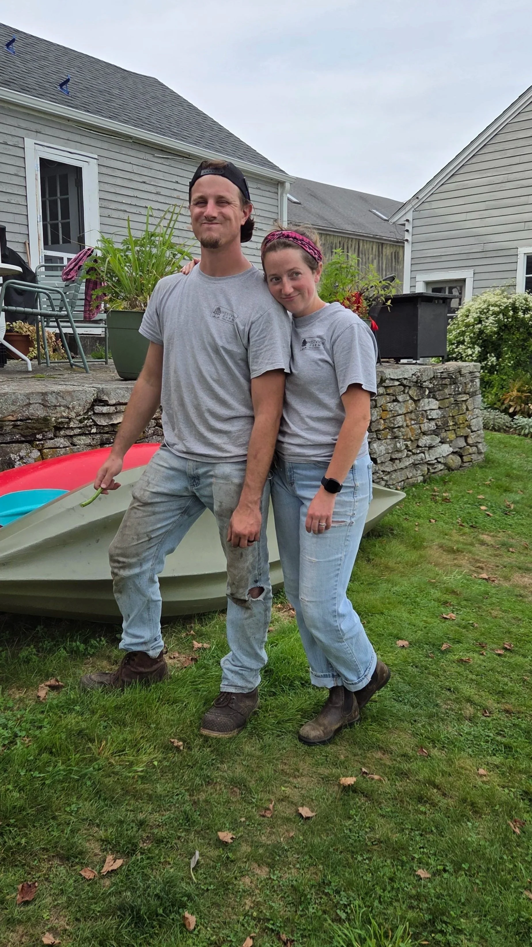 Two people, a man and a woman, standing close together outside on a lawn, smiling. They are dressed casually in gray t-shirts and jeans, with the woman wearing a headband and the man a backward cap. Behind them is a small boat, a stone wall, and a house with potted plants and outdoor furniture.