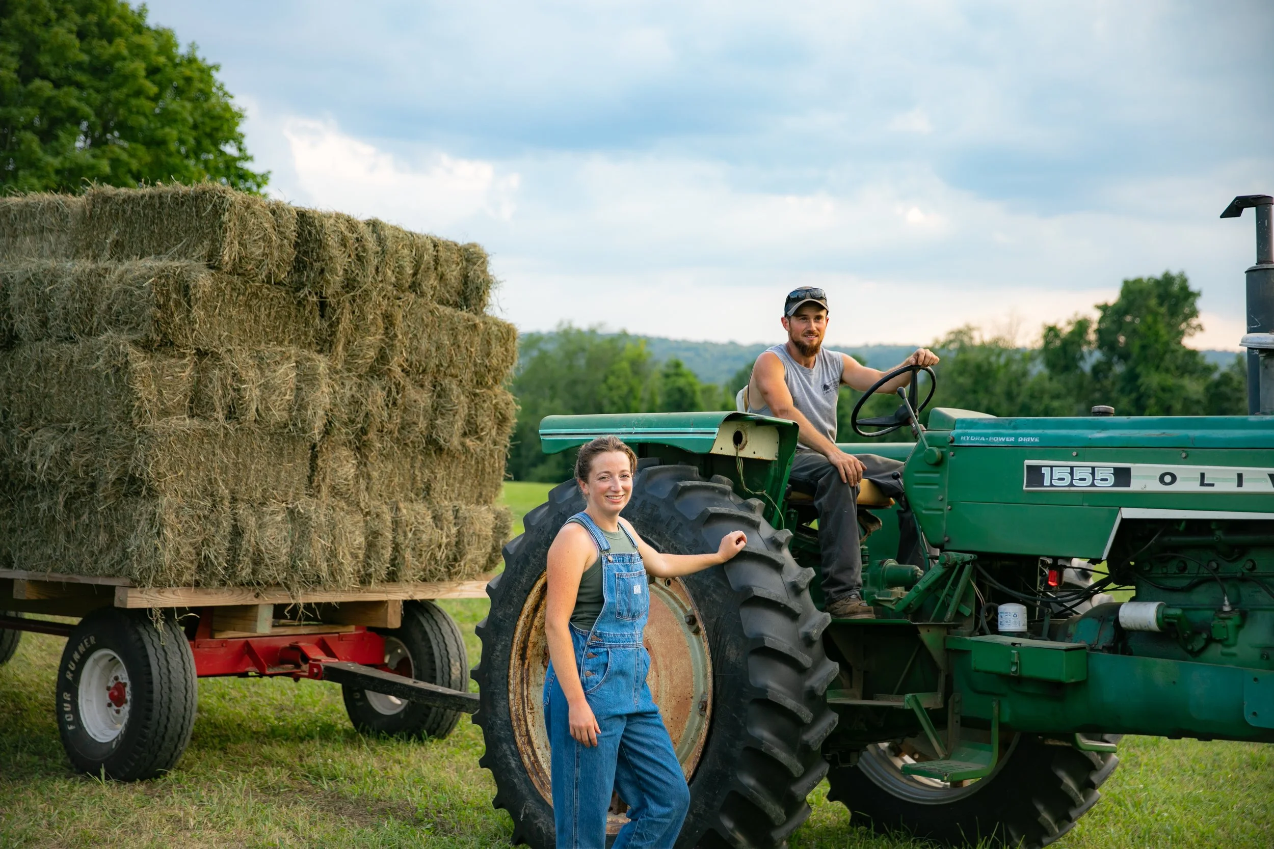 A man and a woman on a farm with a green tractor. The woman stands beside the tractor, smiling, while the man is sitting on the tractor. Behind them, there is a large trailer loaded with hay bales. The scene is set outdoors with green trees and a clo