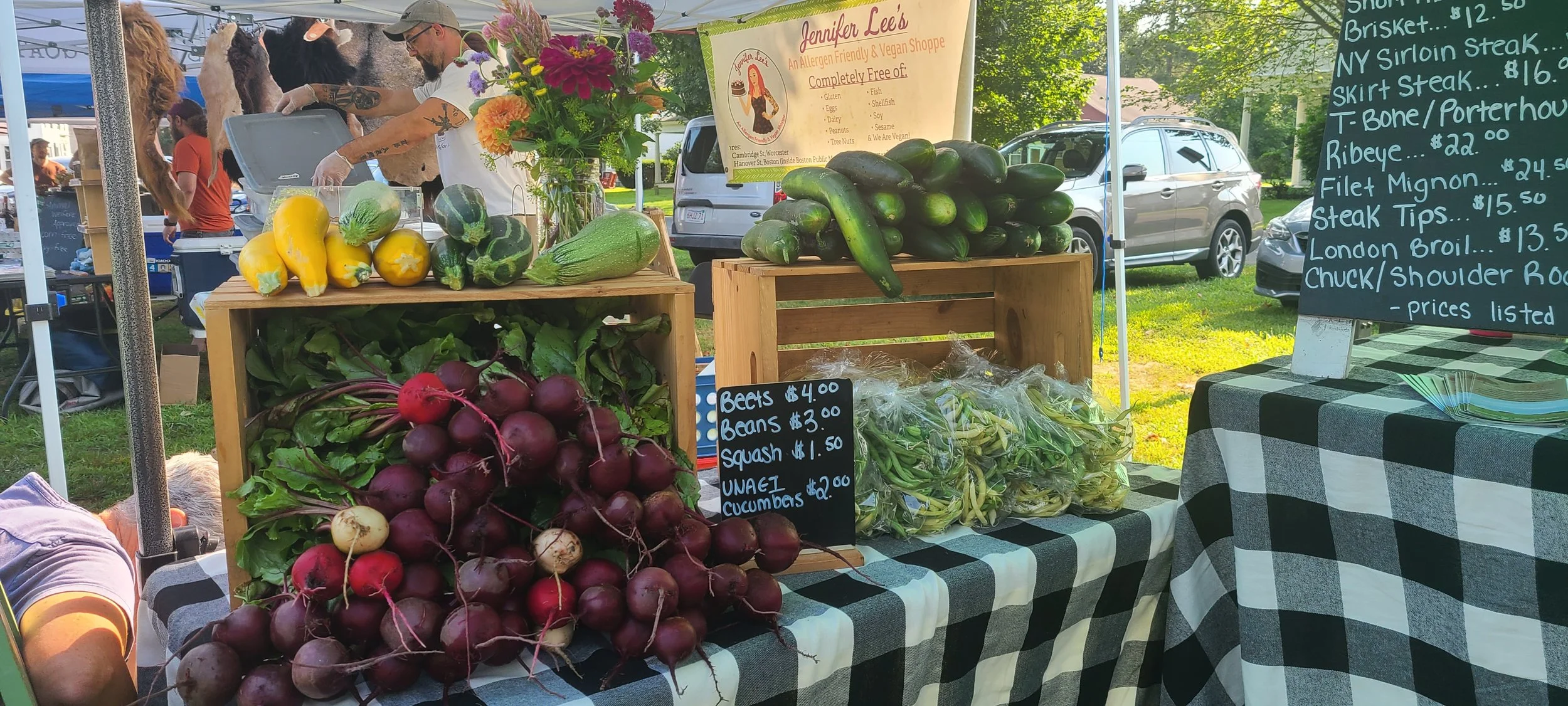 An outdoor farmer's market stall selling fresh vegetables, including beets, squash, zucchini, cucumber, and greens, with a menu sign listing prices and items on a black-and-white checkered tablecloth.