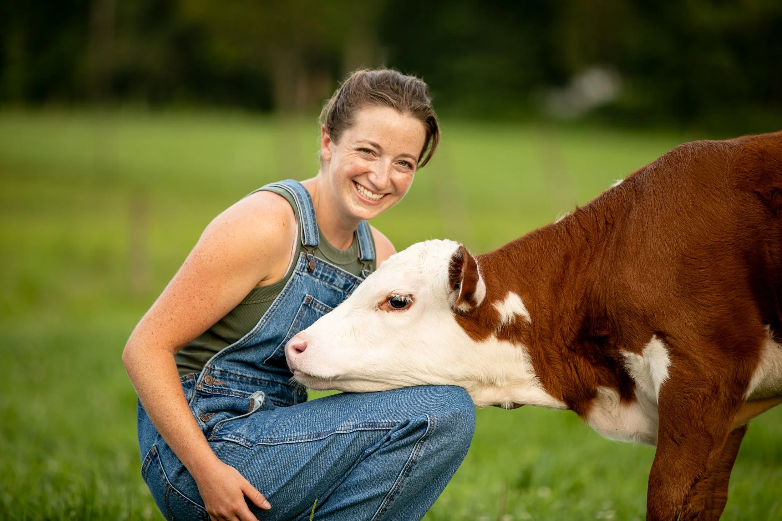 A smiling woman in denim overalls kneeling in a green field with a white and brown calf.
