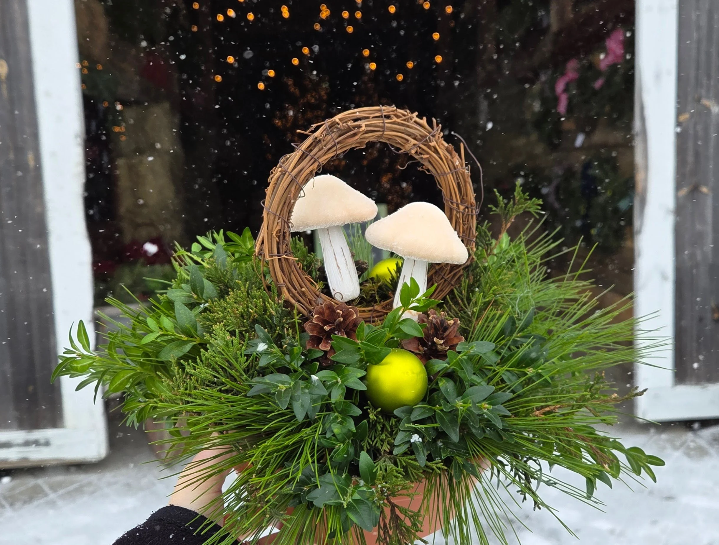 A festive holiday arrangement with two white mushrooms, pinecones, green foliage, and a shiny green ornament, set against a snowy outdoor background.