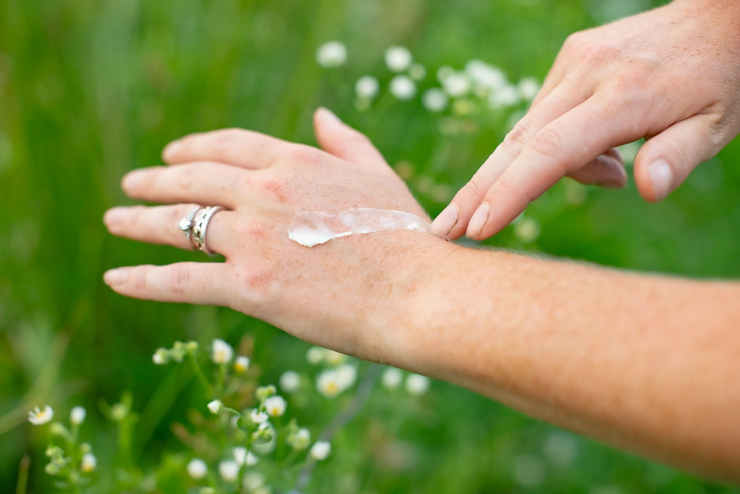 A person applying tallow balm on their hand outdoors amidst greenery and small white flowers.